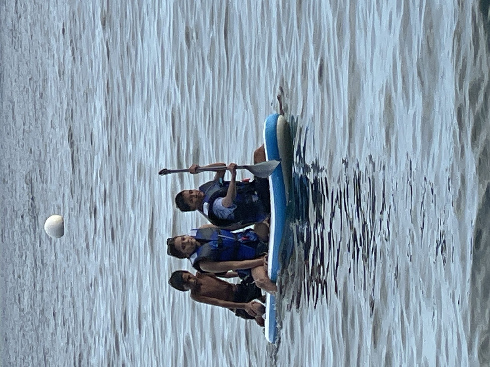 The image shows three young people on a paddleboard in the water, likely near Dauin, Philippines, as indicated by the presence of a spherical buoy floating in the distance, a common sight in coastal areas. The person in the front right is holding a paddle and appears to be in control of the board. They are wearing a blue life vest. To their left, another individual, also wearing a blue life vest, is seated. The third person, on the far left, is kneeling and appears to be shirtless. The water is a muted blue, with visible ripples indicating slight movement. The overall scene captures a moment of recreational activity on the water.