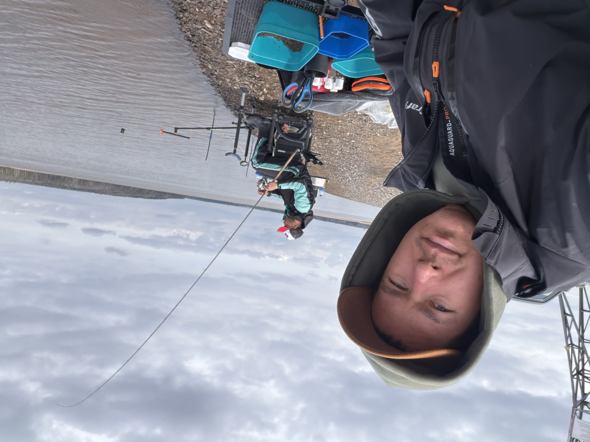 A man is taking a selfie while fishing at the Votkinsk Reservoir in Chaykovsky, Russia. Another person is seen further down the shore sitting in a fishing chair with fishing gear. The man taking the selfie is wearing a hooded jacket and a cap under the hood. The background shows the cloudy sky and the water of the reservoir. Fishing rods are visible, and there's fishing equipment near the seated fisherman.