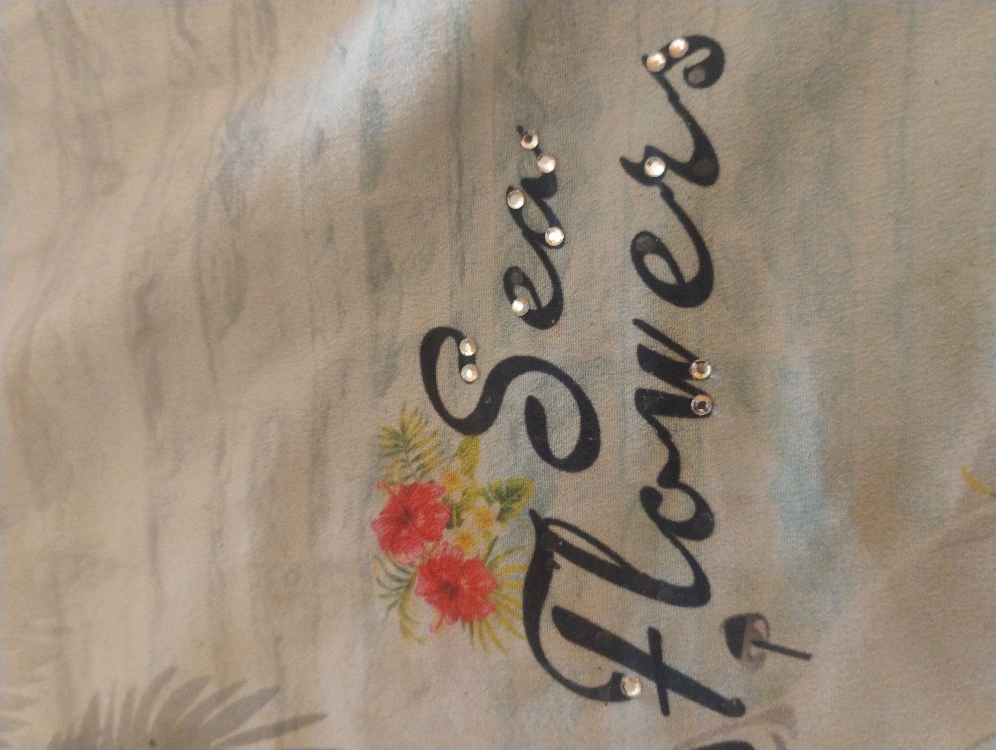 A close-up shot shows a piece of fabric with the words "Sea Flowers" printed in a cursive font. The lettering is black and adorned with small, sparkling rhinestones. To the left of the text, there is an illustration of two red hibiscus flowers with green foliage. The background of the fabric is a light blue-gray with subtle wavy patterns. There is no indication of the location in Rumyantsevo, Bulgaria, or any people present in the image.