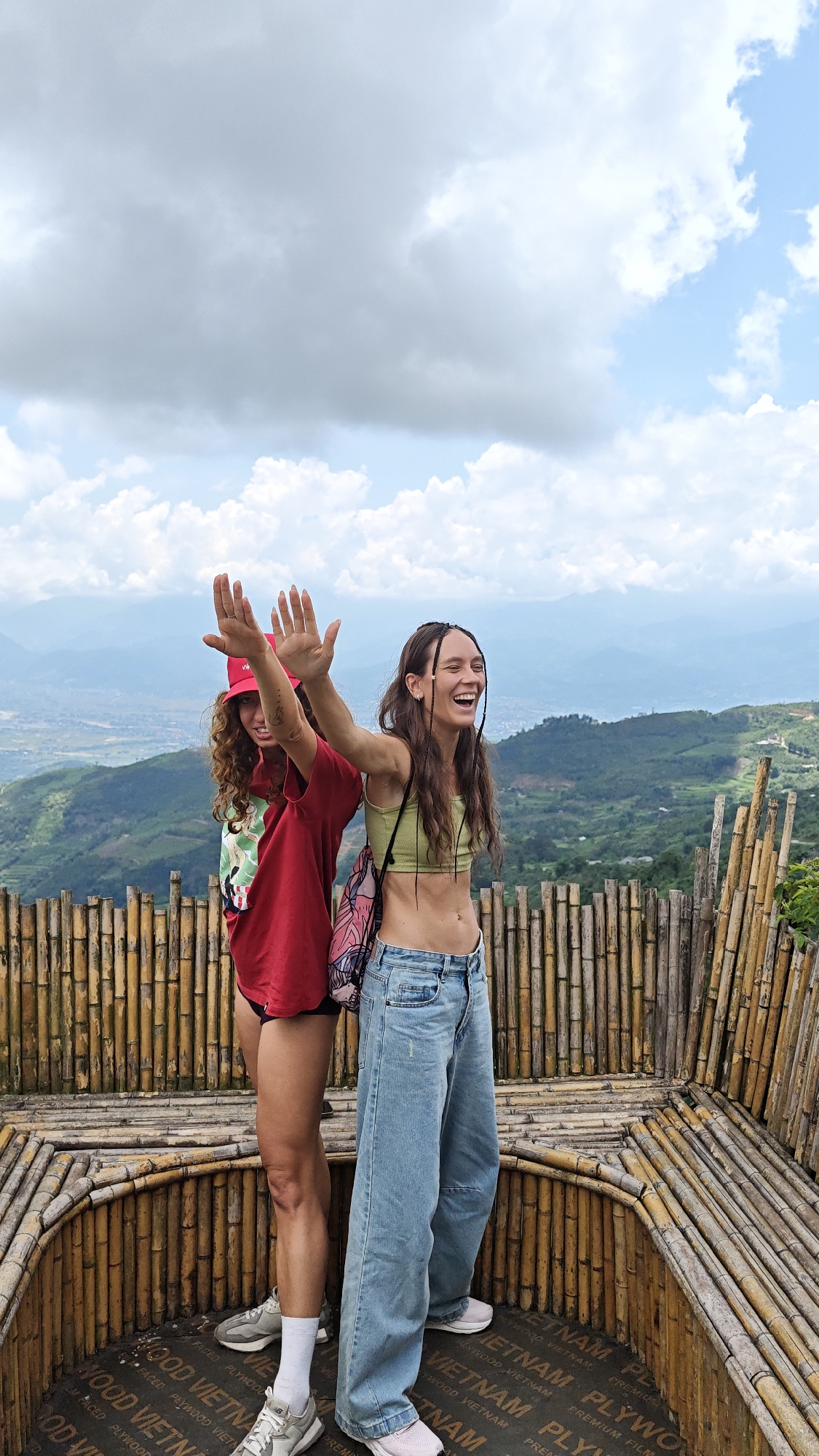 Two young women stand on a bamboo platform, smiling and waving, against a scenic backdrop. The platform is constructed from bamboo poles and rests on a base of plywood, marked with repeated text "WOOD VIETNAM" and "FACED PLYWOOD".
The woman on the left wears a red baseball cap and a red t-shirt. Her hair is curly and she is wearing white sneakers. The woman on the right has long brown hair with dark braids woven into it. She wears a green cropped top and loose, light-blue jeans. She has a pink and blue bag slung over her shoulder.
The platform overlooks a landscape of green hills, mountains, and a valley in the distance, partially obscured by clouds. The sky is overcast with a mix of dark and light clouds. The scene is located in or near Suối Giàng, Vietnam.