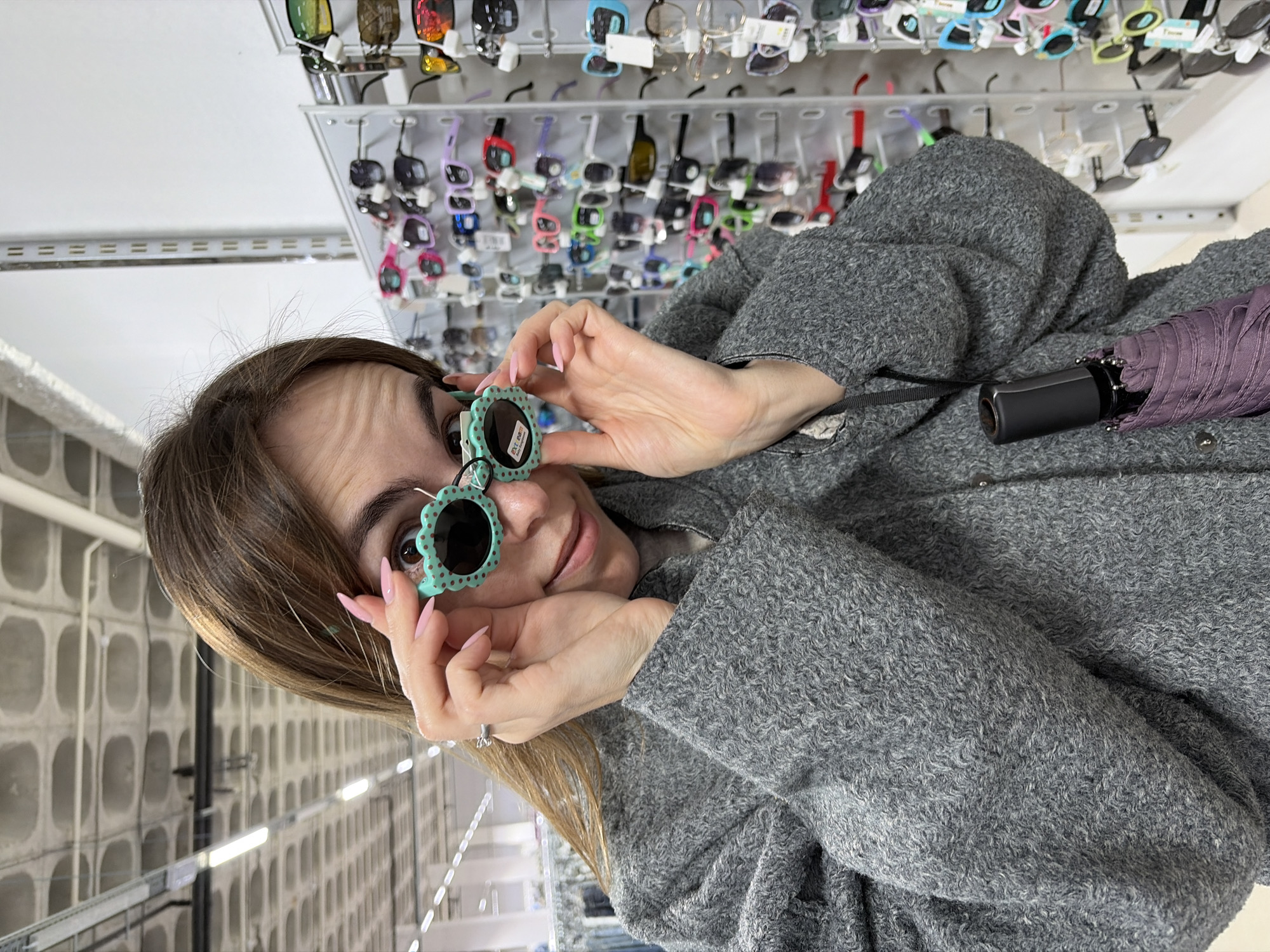 The original image is rotated 90 degrees clockwise. It depicts a young woman in an indoor retail setting, likely a sunglasses store or a market stall in Izhevsk, Russia.
**Subjects:** The primary subject is a young woman with brown hair, shown from the chest up, looking directly at the camera. Her face shows a slight, playful smile, and her eyebrows are raised, creating lines on her forehead. She has light pink manicured nails and is wearing a ring on her left ring finger. She is dressed in a textured, curly grey jacket or coat. A closed purple umbrella with a black handle hangs from her shoulder or arm.
**Objects:** The woman is holding a pair of novelty, flower-shaped sunglasses to her eyes. These sunglasses have mint green frames with scalloped edges resembling petals and dark, round lenses. Behind her, multiple shelves are filled with various sunglasses of different styles and colors, hung on hooks or displayed on racks. A small, illegible white tag is visible on the arm of the flower sunglasses.
**Scene and Setting:** The background immediately behind the woman is a display area with numerous sunglasses. Further back, the scene transitions to an industrial-looking interior space characterized by large, grey concrete walls with a grid pattern of recessed square or rectangular indentations. Fluorescent light fixtures illuminate this background area, suggesting a large building or warehouse-like environment within the retail complex.
**Activity:** The woman is engaged in the act of trying on or posing with a whimsical pair of sunglasses, possibly deciding on a purchase or simply having fun in the store. Her direct gaze at the camera suggests she is being photographed while doing so.
**Notable Details:** The image's rotated orientation gives it an unusual perspective. The woman's expression is one of lightheartedness or amusement. The varied sunglasses on display highlight the retail nature of the location, while the stark concrete background provides an interesting contrast to the colorful products and playful activity. No clearly legible text is visible on any of the objects or in the background.