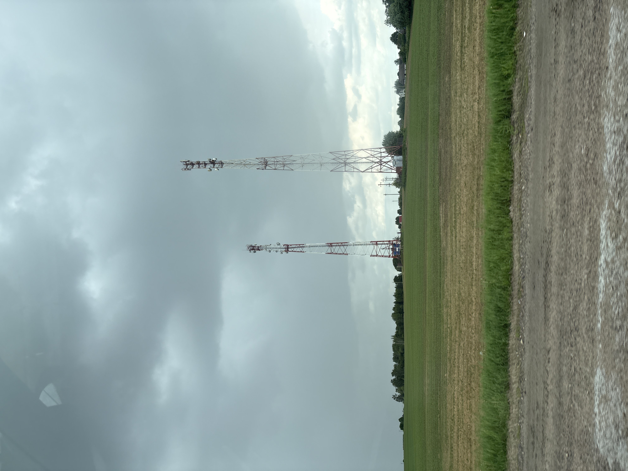 The image depicts a rural landscape in Голацкі сельскі Савет, Belarus. Two communication towers stand prominently against a cloudy, overcast sky. The towers are red and white lattice structures. A field of green and brown grass extends to the right of the towers. A dirt or gravel road runs along the right edge of the image. Trees and vegetation are visible in the distance. The light suggests it is daytime.