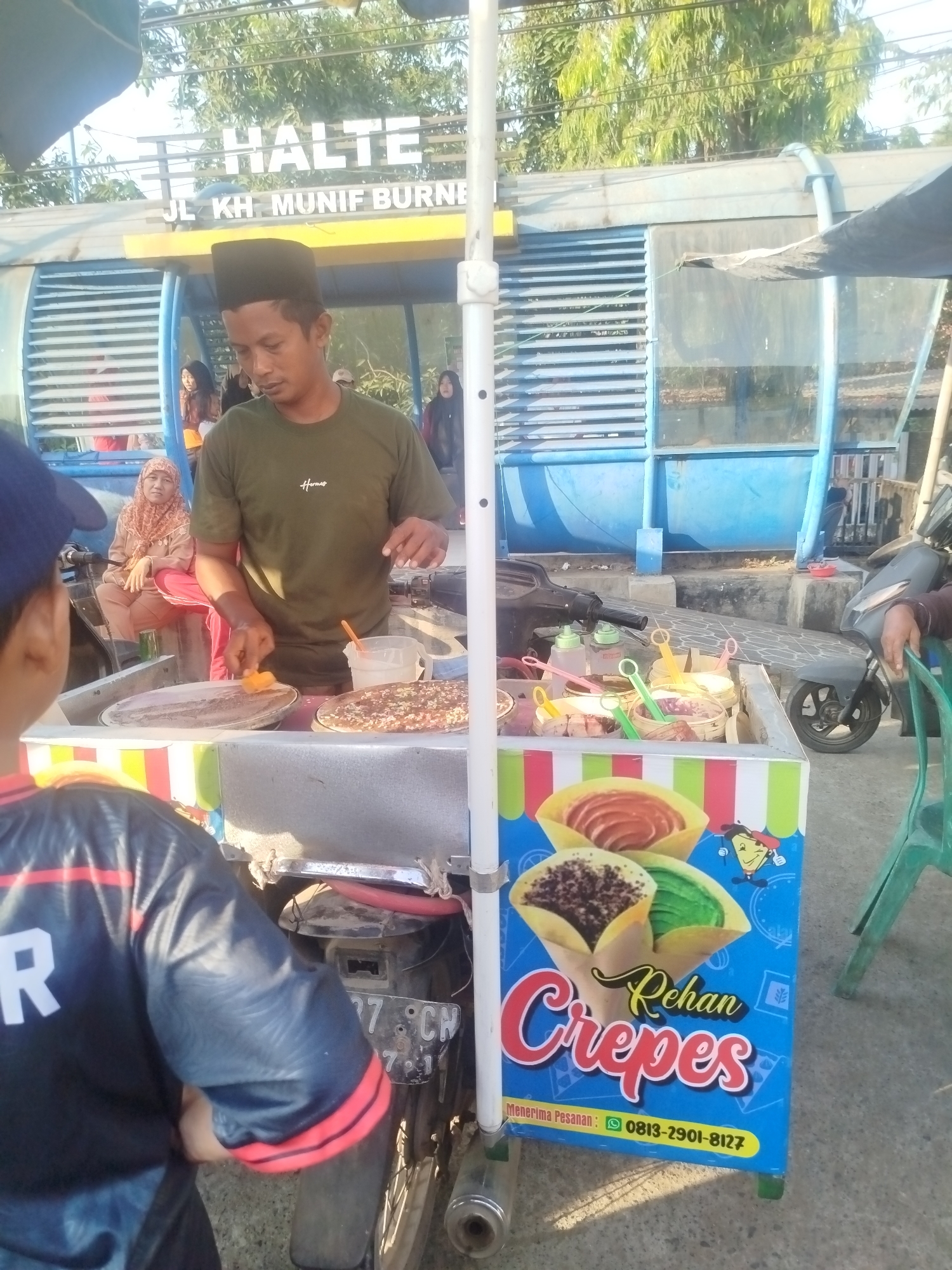 In Burneh, Indonesia, a street vendor is preparing crepes at a cart situated near a bus stop identified by the sign "HALTE JL. KH. MUNIF BURNE." The vendor, a man wearing a dark cap and a green t-shirt, is actively cooking on a large, flat griddle. He is holding a scraper tool in his right hand, mid-action on a crepe.
The crepe cart is adorned with a colorful banner displaying images of crepes and the text "Rehan Crepes" along with a phone number. Inside the cart, various toppings and ingredients are visible in separate containers, some with green and pink serving spoons. Behind the vendor, a woman in a patterned hijab is seated, observing the scene. A young person wearing a blue cap and a black and red shirt is positioned in the foreground, looking towards the vendor.
The background features a blue bus stop structure with slatted windows and the surrounding environment includes trees, a sidewalk, and a parked motorcycle. The overall scene depicts a typical street food vending operation in an urban setting.
