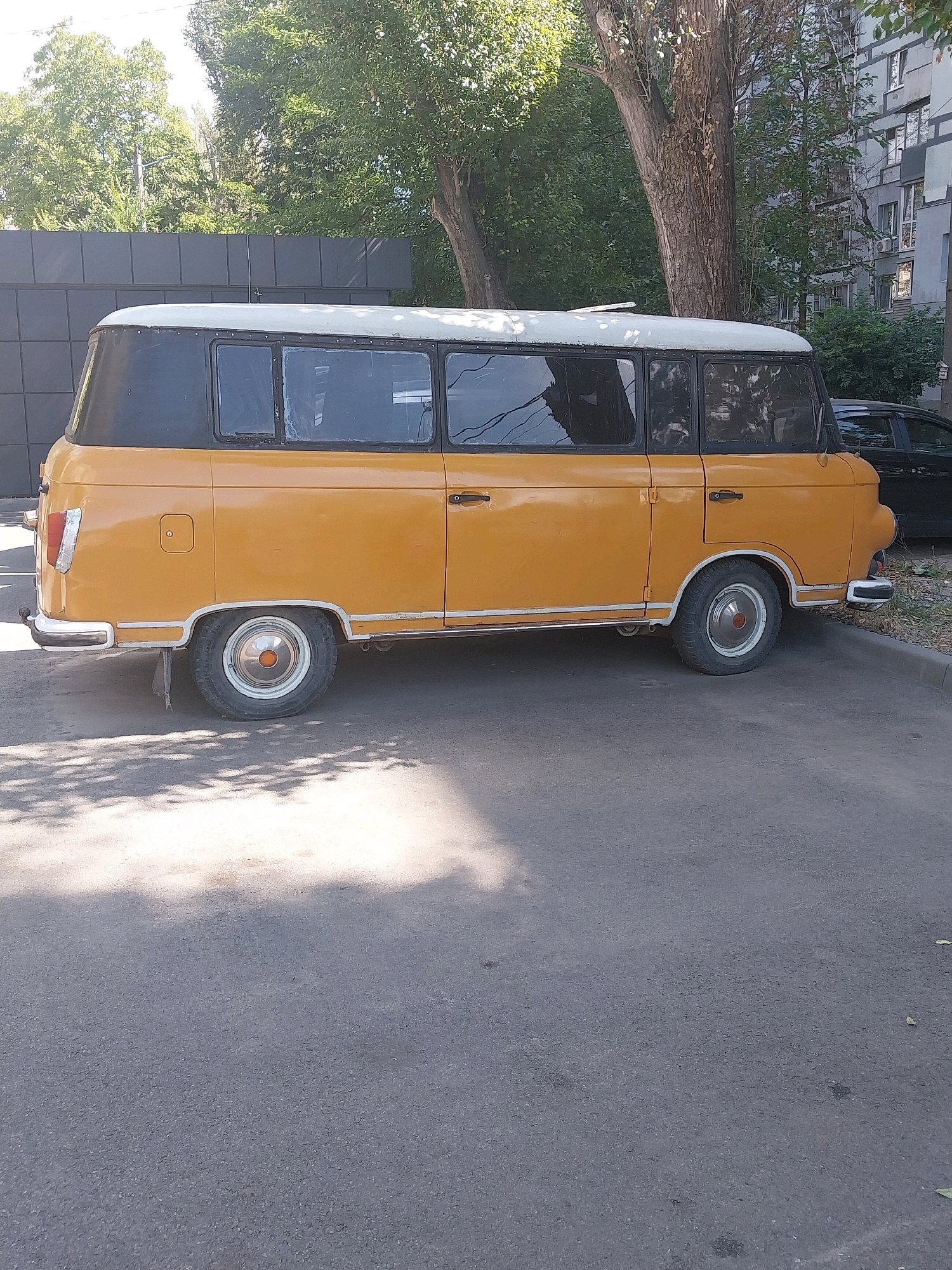 A vintage yellow van, possibly a UAZ-452, is parked on an asphalt surface in a city. The van has a distinctive two-tone color scheme with a black roof and grey trim around the windows. It appears to be a passenger van with multiple windows. In the background, there is a building with multiple stories and windows, suggesting an urban environment. Trees with green foliage are also visible, casting shadows on the ground. The scene is lit by sunlight, creating bright highlights and shadows. The van is the primary subject, and it is stationary. The image captures an outdoor scene during daylight hours. The location is identified as Dnipro, Ukraine.