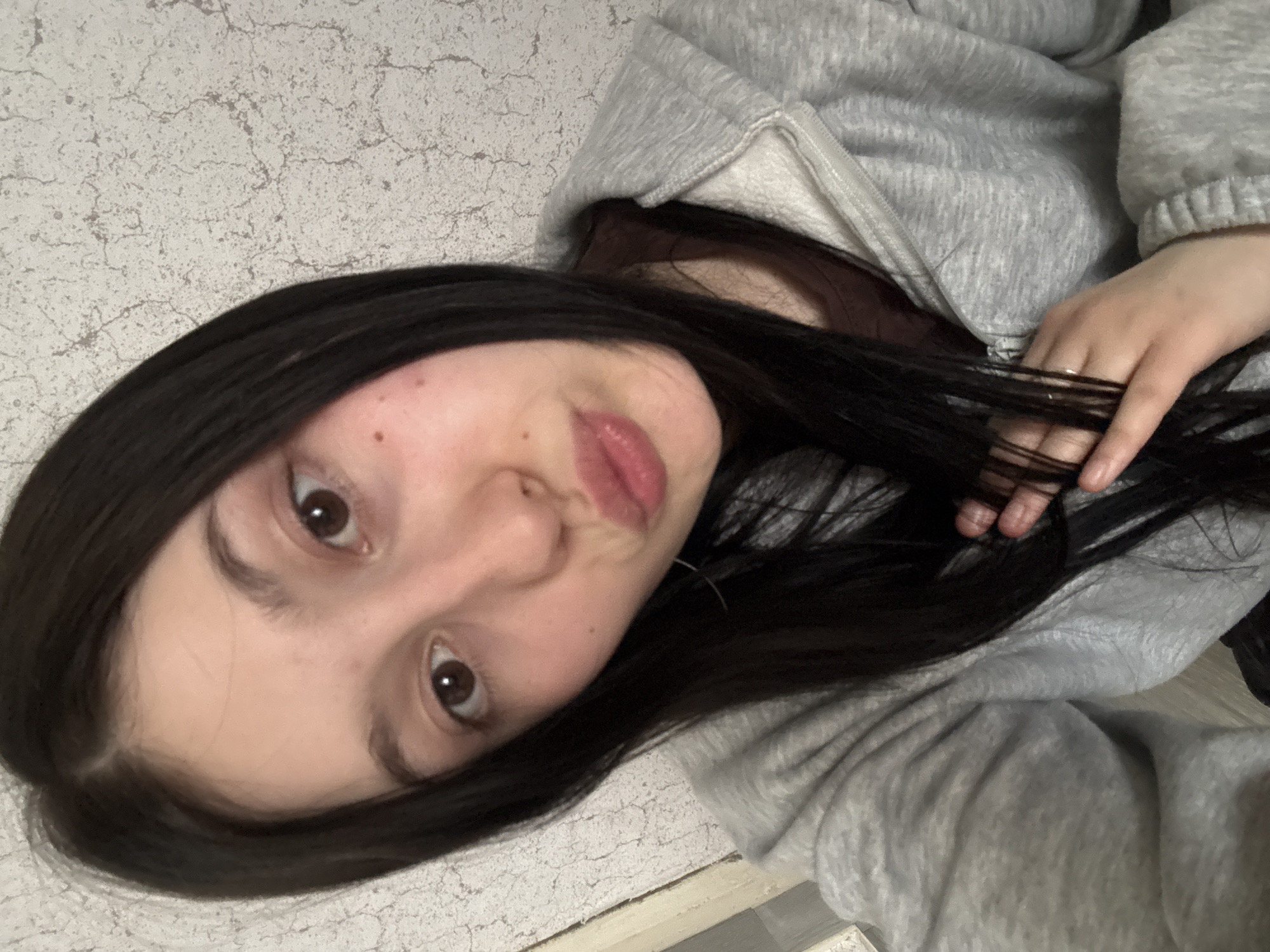 A young person with fair skin, brown eyes, and dark hair is posing for a selfie indoors. They are wearing a light gray hooded sweatshirt. The background appears to be a wall with a light-colored, crackled or textured surface. The location is unknown and cannot be determined to be in the city of Фоки, Russia.