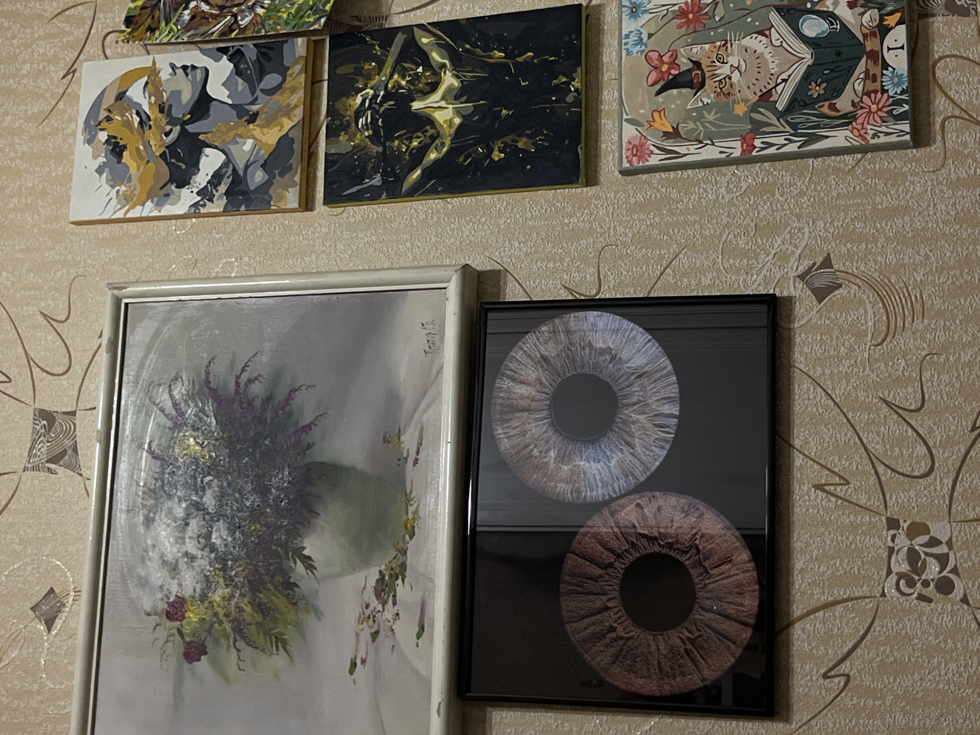 The image shows a section of a wall with patterned wallpaper. Four framed pictures hang on the wall. The top row includes two small square abstract paintings and a painting depicting a cat reading a book. The bottom row features a framed painting of flowers and a framed image of two irises. There is no information in the image to confirm the location is Rostov-on-Don, Russia.