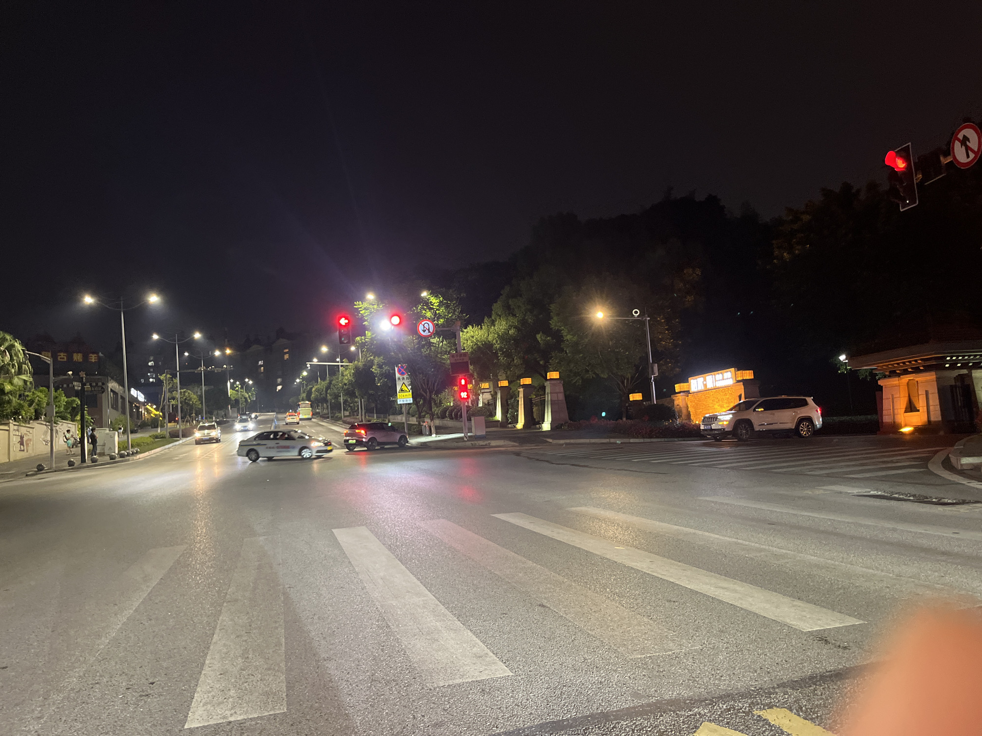 The image depicts a nighttime street scene in Jiulongpo District, China, focusing on a road intersection with a pedestrian crossing. Several cars are visible on the asphalt road, some illuminated by streetlights and traffic signals, which display red lights, indicating a stop. On the left side of the road, there are buildings with illuminated signage and decorative elements. Trees and streetlights line the sidewalks. The sky is dark, typical of nighttime, and distant hills are faintly visible in the background. The scene appears to be a moderately busy urban area at night.