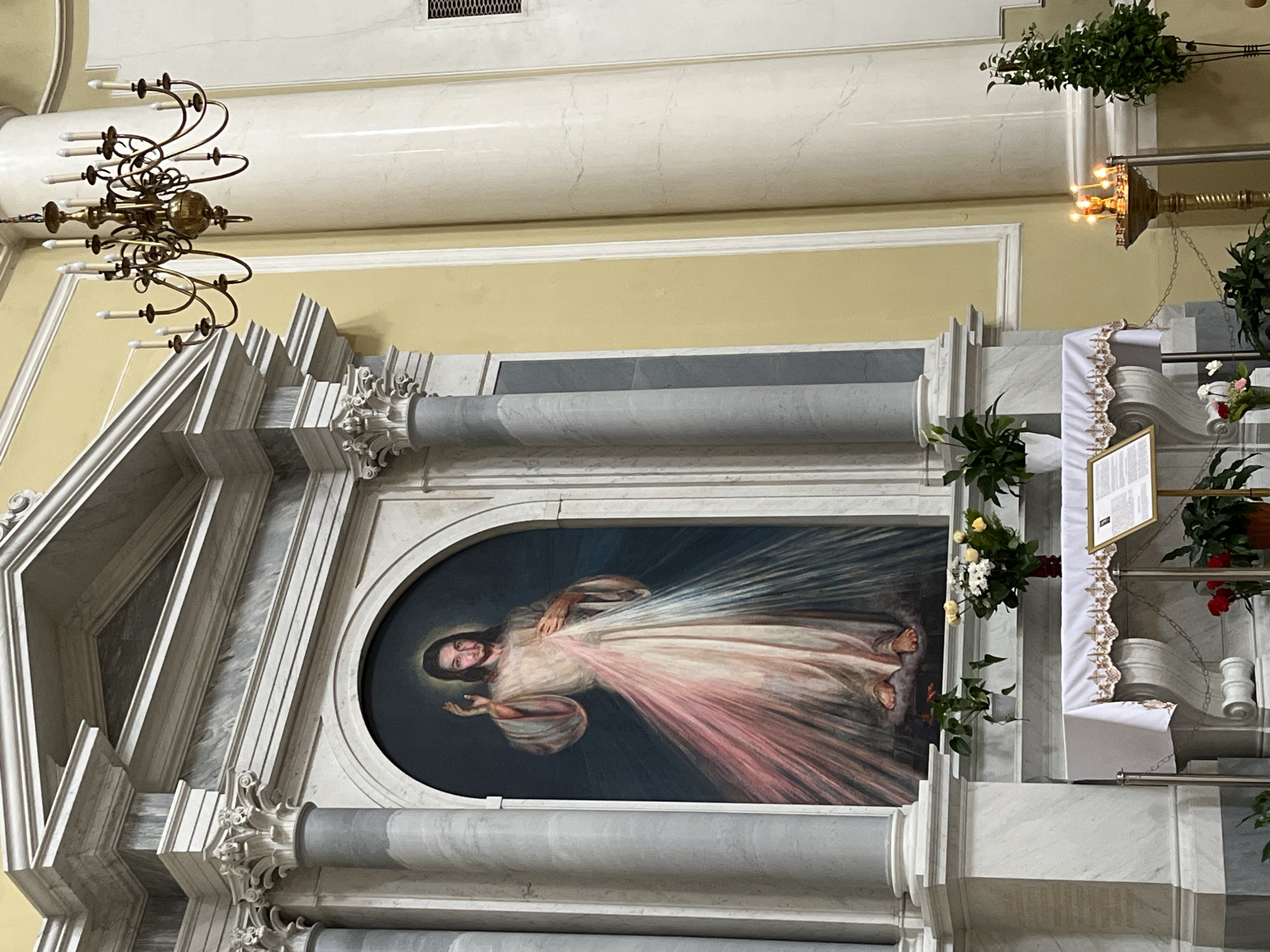 The image shows the interior of a church. In the center is a painting of Jesus Christ, with rays emanating from his heart. The painting is set within an elaborate marble frame with columns and decorative elements. To the right of the painting is a display area with flowers, a draped cloth, and what appears to be a sign or notice. In the upper-left corner, a brass chandelier hangs from the ceiling. The walls are painted a light yellow color, and the architecture suggests a religious building. The location is Saint Petersburg, Russia, based on the architectural style and likely religious denomination.
