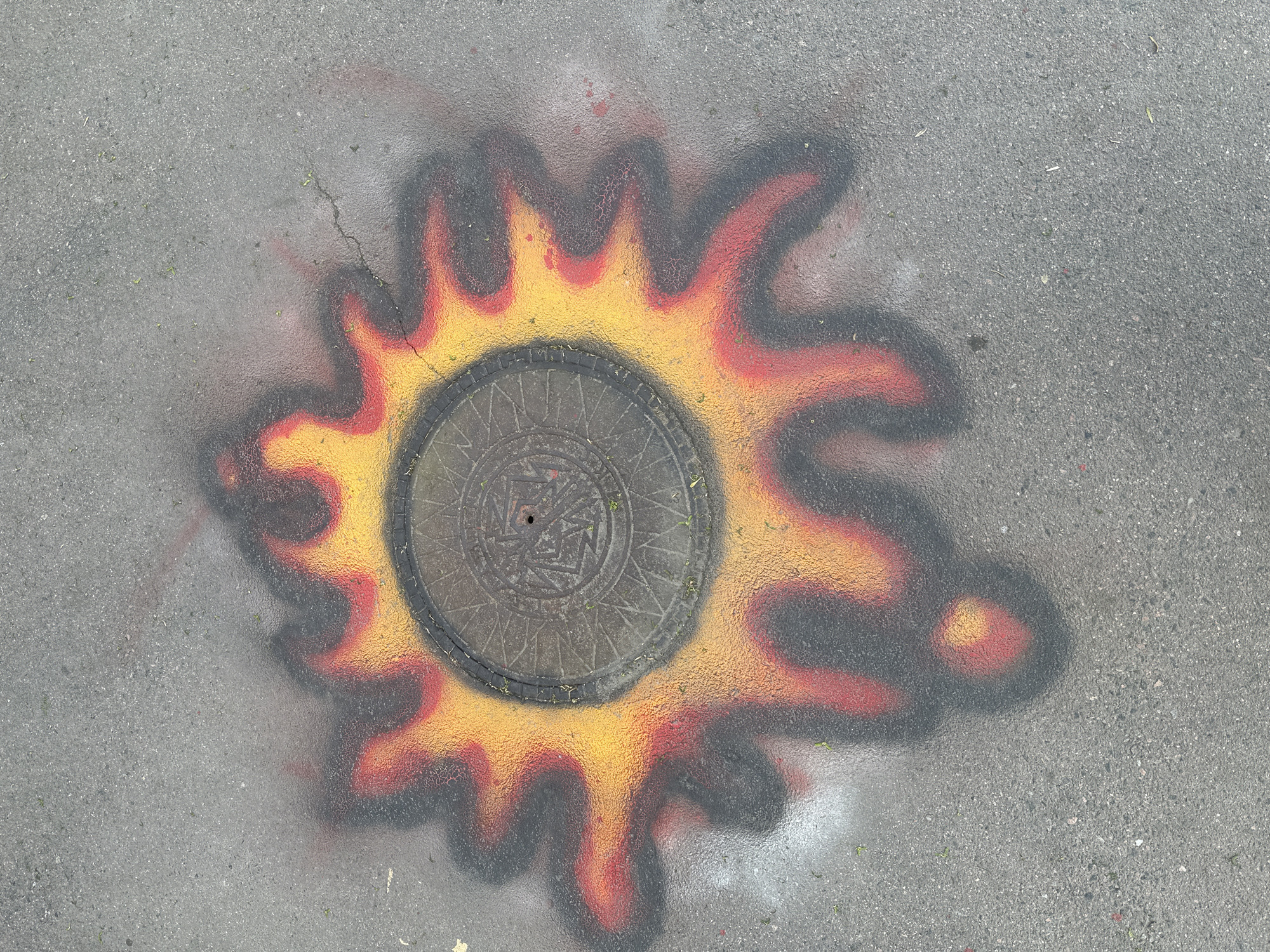 A manhole cover is spray-painted to resemble a sun or flame on a gray asphalt surface. The central manhole cover is a dull gray with a circular pattern and a small hole. The painted design surrounding the manhole consists of yellow, red, and black concentric layers, mimicking the appearance of flames or the sun's corona. The asphalt surface is textured and shows signs of wear, with minor cracks visible near the painted area. The image does not contain people. The location in the city of Saint Petersburg, Russia, is not discernible from the image.