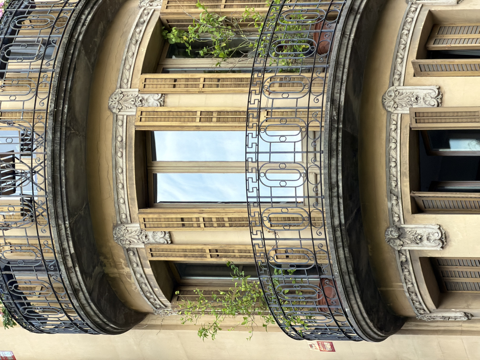 The original image, rotated 90 degrees clockwise, and its crops display a detailed section of an ornate building facade, likely located in Barcelona, Spain. The multi-story structure features several curved balconies, each adorned with intricate black wrought iron railings showcasing repetitive oval and geometric patterns. The facade itself is a light, warm beige color, intricately decorated with carved floral motifs and classical moldings around the windows and supporting the balconies. Many windows are equipped with light-colored wooden louvers or shutters, some of which are open while others are closed. The bright, clear sky is reflected in several windowpanes, indicating a sunny daytime scene. Green plants, some appearing to be in terracotta pots, spill over the railings of certain balconies. A red and white sign displaying the word "Alarma" is visible on the lower left side of the original image, attached to the building near a balcony. No people or specific events are depicted; the focus is solely on the architectural details.