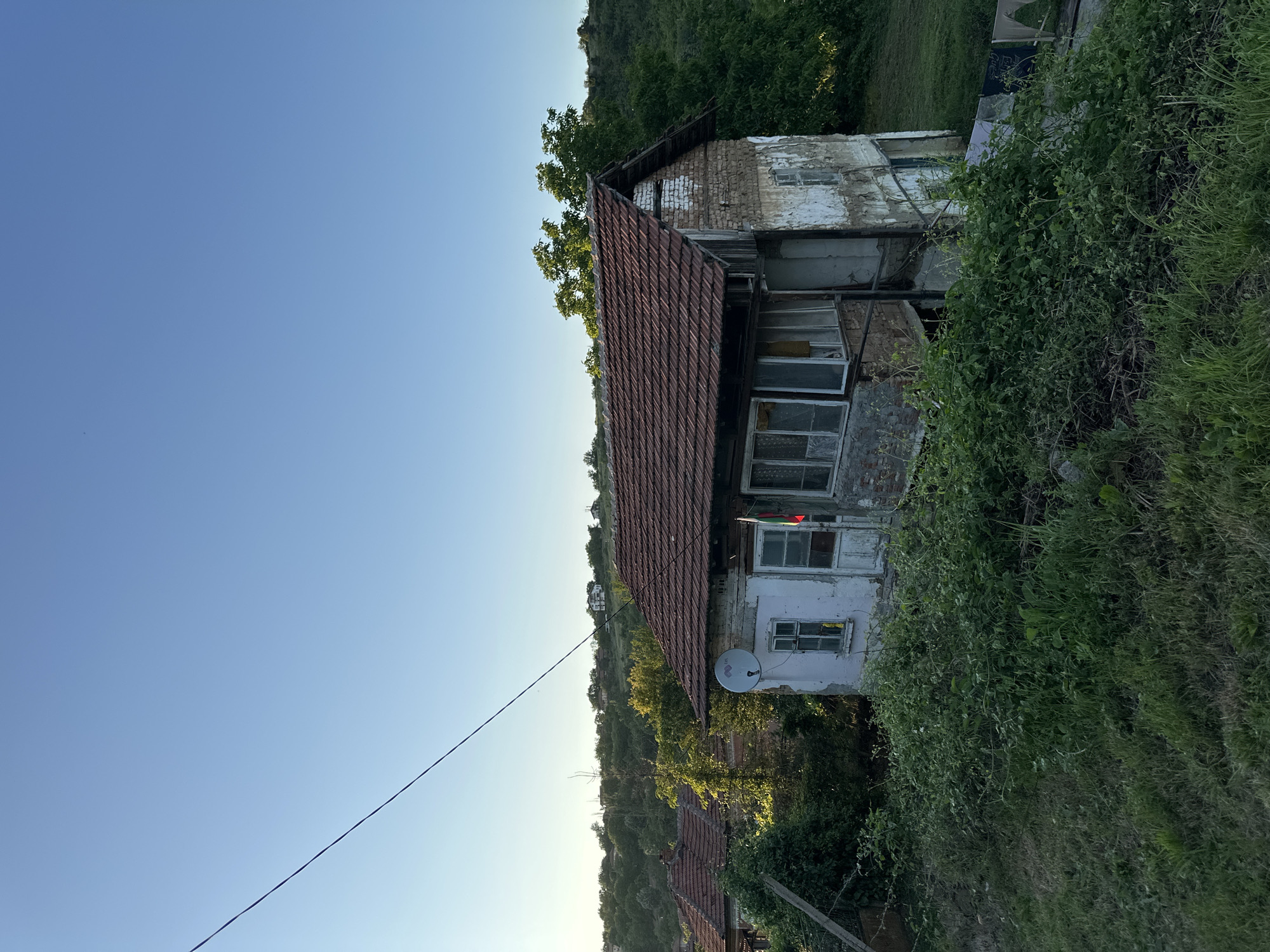 The image depicts an aged, two-story house nestled on a verdant hillside, characteristic of a rural Bulgarian village like Slavyani. The house features a distinctive reddish-brown tiled roof and walls that display a blend of peeling white plaster, exposed brick, and older construction, indicating significant wear. Several windows are visible, some appearing weathered or damaged, with a small flag, possibly red and green, hanging near an upper window on the right. A white circular satellite dish is affixed to the front facade below a window.
The property is enveloped by lush, overgrown green vegetation, including thick bushes, vines, and tall grasses, especially prominent on the right side. Beyond the immediate foreground, rolling hills densely covered in trees stretch into the distance, with the roof of another dwelling peeking out further down the slope. The sky is a clear, bright blue, suggesting fair weather and daylight, possibly late afternoon, given the quality of light. A solitary power line extends diagonally across the upper left portion of the serene landscape. No people are present, and no specific activities are underway.