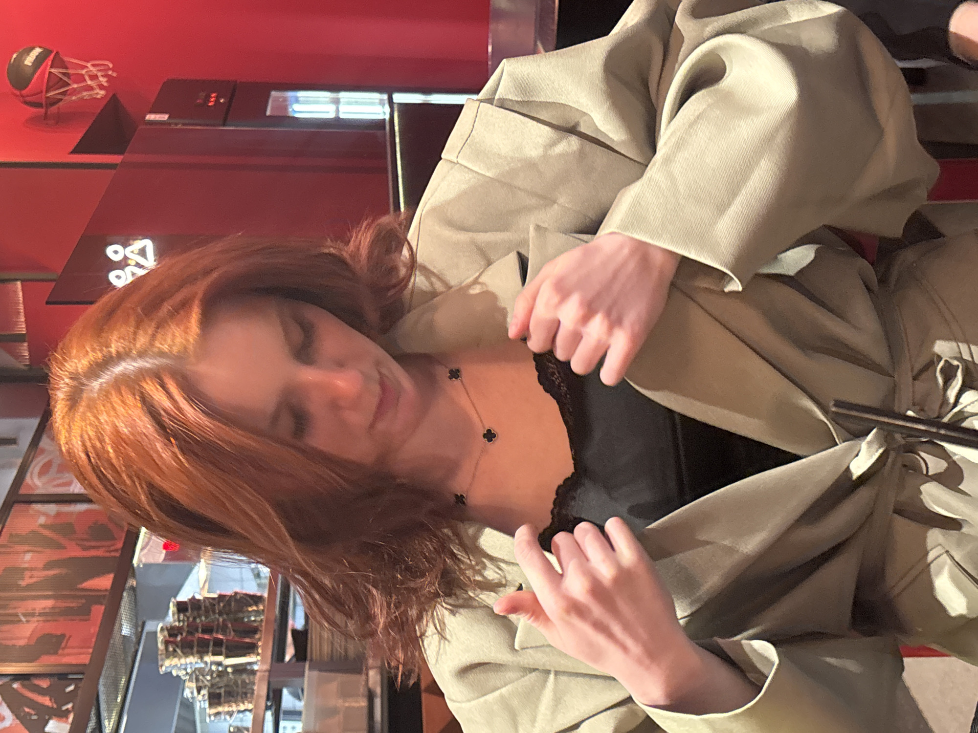 A medium indoor shot depicts a young woman with shoulder-length red hair, wearing a black top and a beige trench coat. She is adjusting the lapels of the coat with both hands, looking down slightly. A black necklace with a clover-leaf design adorns her neck. In the background, there is a red wall with decorative elements, including a miniature basketball hoop. The scene is set in a restaurant or bar with reflective glass, but the exact location in the city of Unknown, Russia is not discernible from the image.