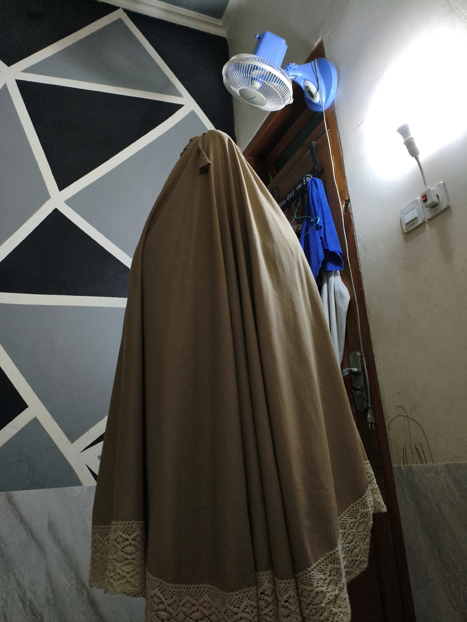 An indoor, low-angle, medium shot shows a person wearing a tan religious garment that covers their entire body, including their head. The garment features decorative lace trim along the bottom hem. The person stands in front of a wall with a geometric pattern of black, gray, and white triangles. To the right, a wooden doorframe is visible, with a blue fan mounted on the ceiling above it. A bright light illuminates the upper right portion of the scene, emanating from a light bulb plugged into a white electrical outlet. Several items, including a blue shirt and a gray garment, hang on the doorframe. A portion of a marble surface is visible in the lower left and right corners of the image. The overall scene appears to be an interior domestic space within Karangtalun, Indonesia, possibly a bedroom or prayer area.