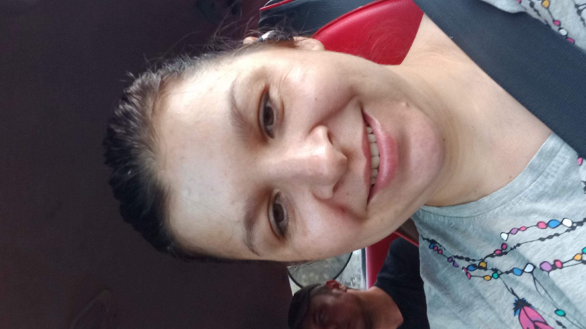 A woman is taking a selfie inside a vehicle. She is in the foreground, smiling and looking at the camera. She has dark hair pulled back and is wearing a gray t-shirt with a colorful beaded pattern. A seatbelt is visible across her chest. In the background, to the left, a man wearing a dark baseball cap and a black t-shirt is visible, partially out of focus. The interior of the car appears to be red and black. The scene is indoors, likely within a car, and does not provide specific details that can pinpoint a location within Kropyvnytskyi, Ukraine.