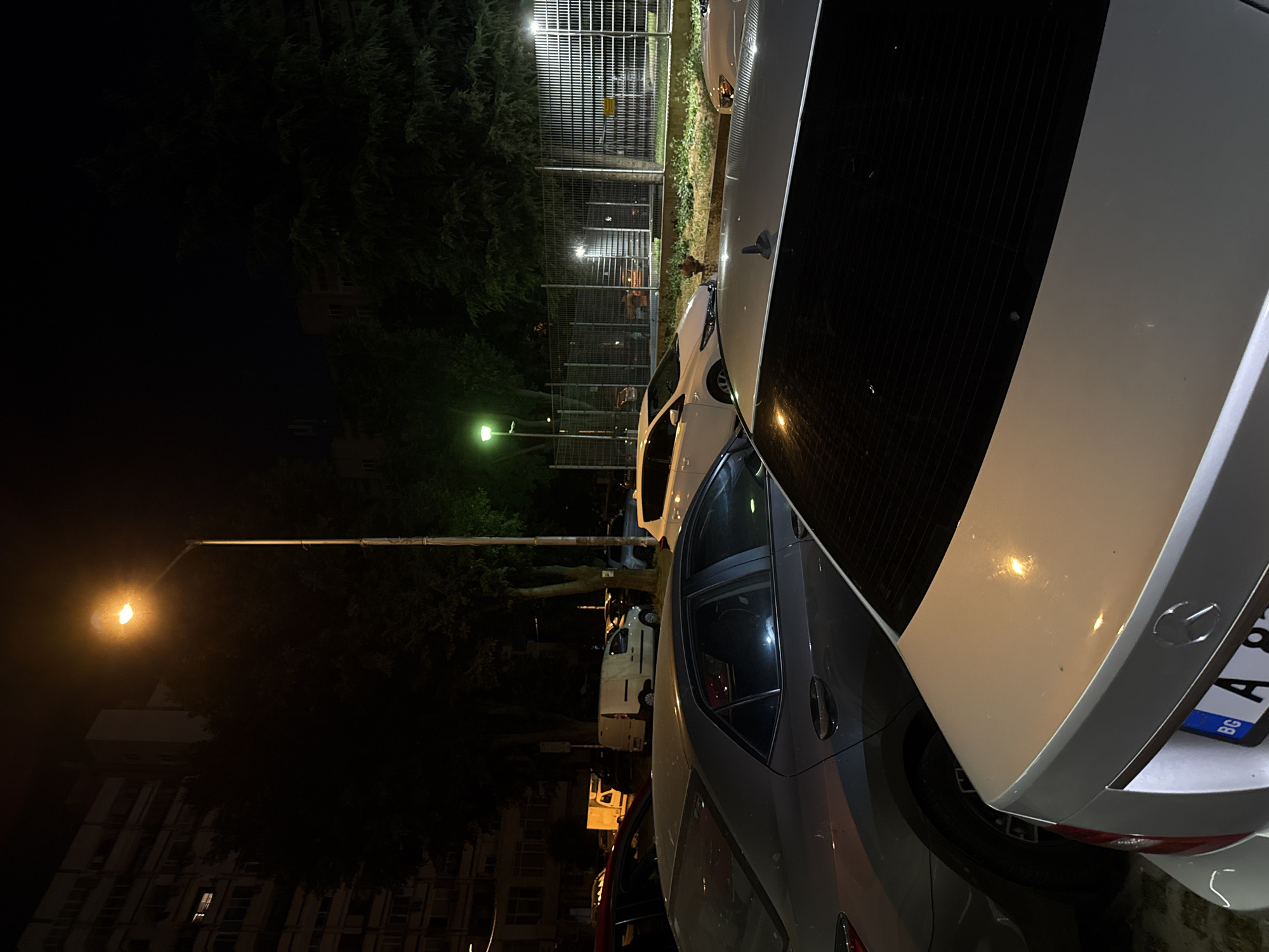 At night, a street lamp illuminates a parking lot filled with several cars. In the foreground, the rear of a silver Mercedes-Benz is visible, with a Bulgarian license plate. To the left, a gray sedan is parked, and further back, a white panel van is parked alongside other vehicles. The background shows a building complex and trees, with a metal fence and more parked cars visible beyond the trees.