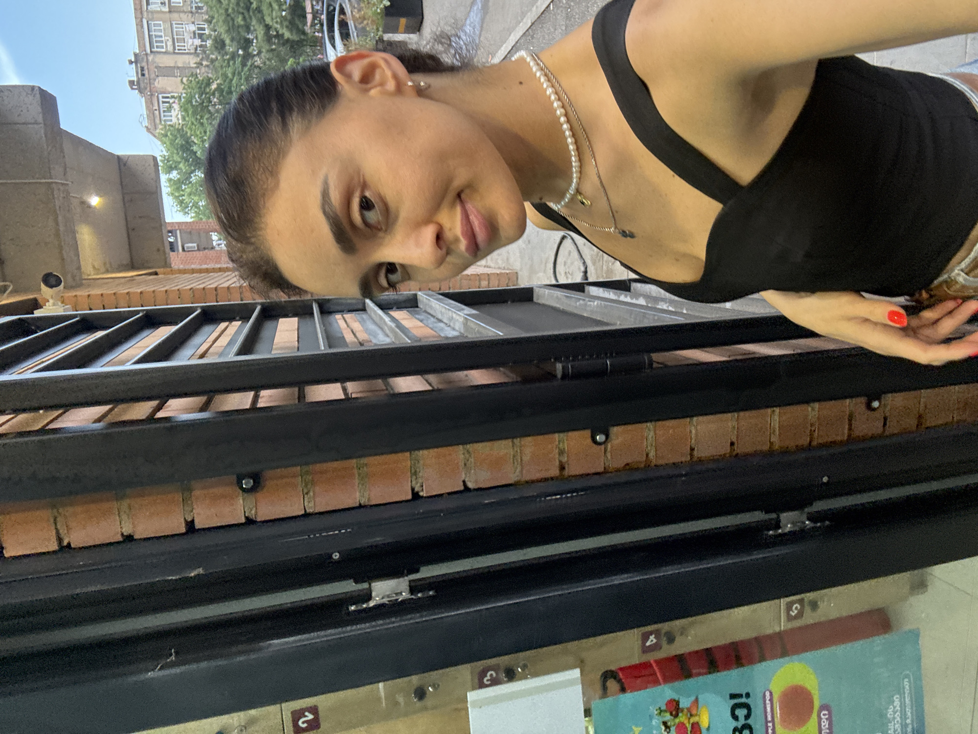 A young woman with dark hair pulled back and a black tank top is taking a selfie outdoors. She is wearing a pearl necklace and a smaller chain necklace. She has red nail polish on her right hand. Behind her, a brick building and a metal grate are visible. The environment suggests an urban setting, possibly a storefront or an entranceway. The presence of Georgian text on a sign to her left strongly indicates the location is Tbilisi, Georgia. The building appears to be a multi-story residential structure. The lighting suggests it is daytime.