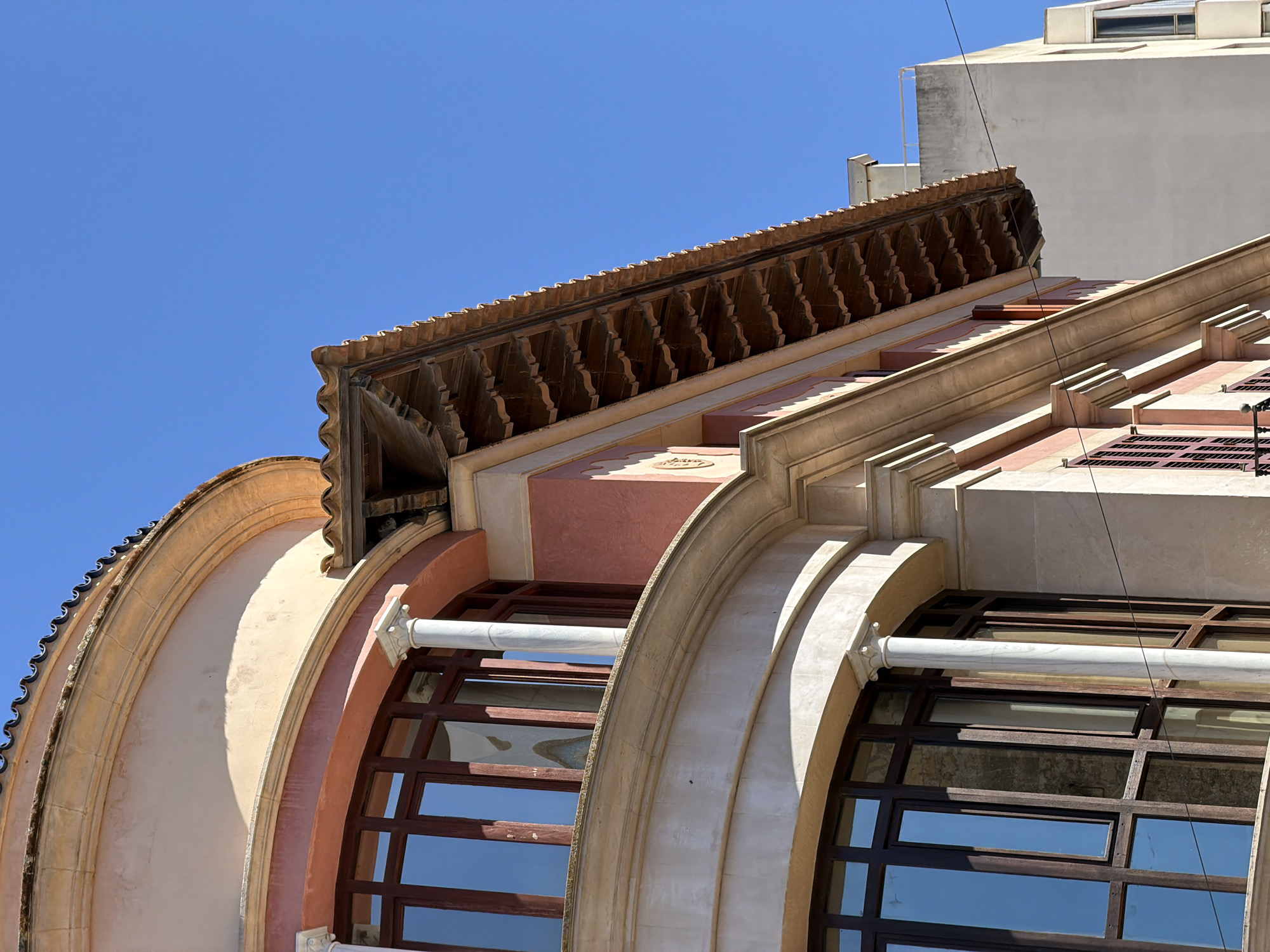 An upward angle shot shows the terracotta tiled roof of a building in Manacor, Spain. The building is painted pink and beige. The roof has a decorative trim. Adjacent to it is a modern building that is light grey. The sky is clear and blue. There are no people or animals visible in the image.