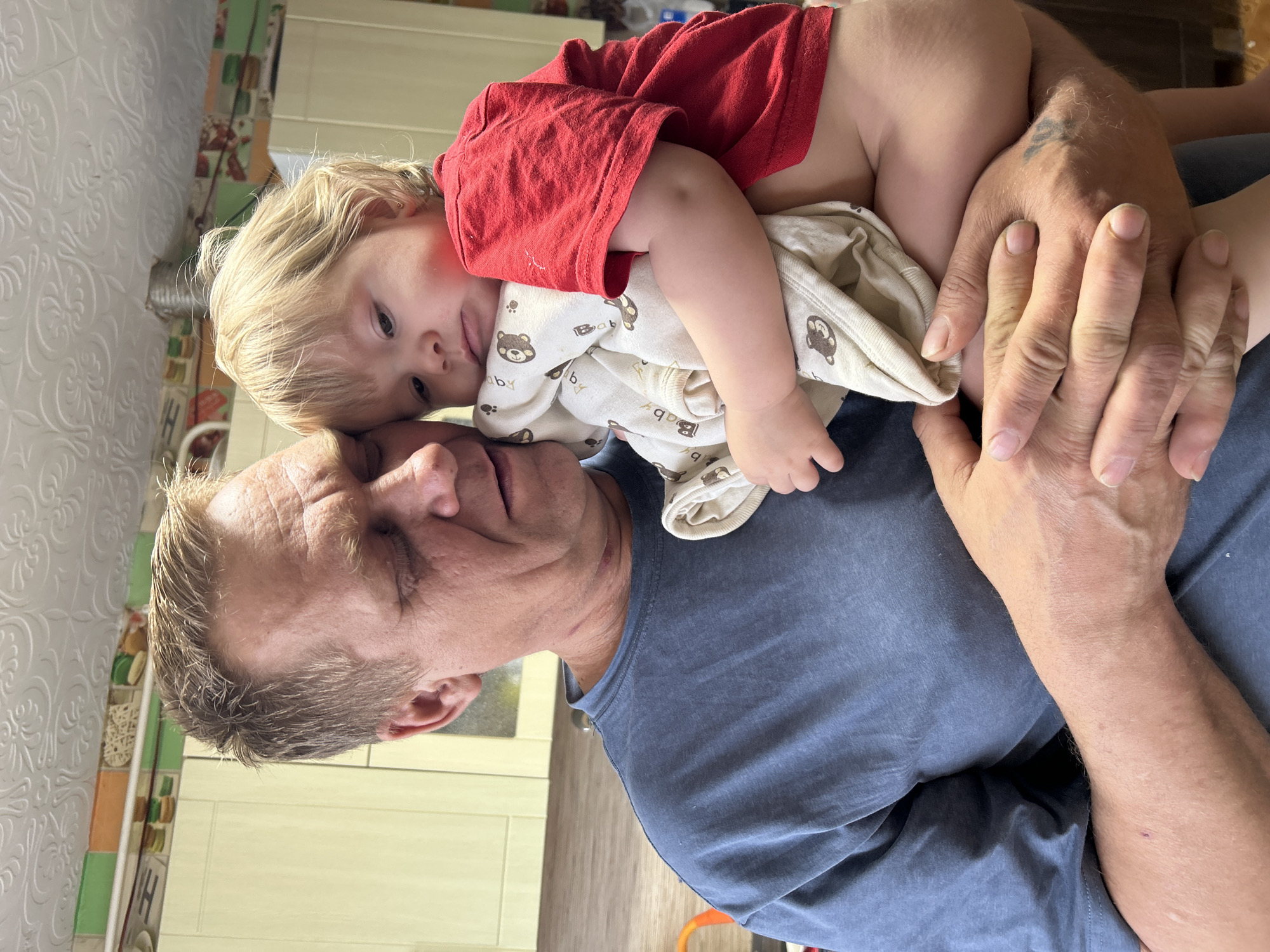 A man with short, light-colored hair and a blue t-shirt is holding a young child. The child, with light-colored hair, is wearing a red t-shirt and a cream-colored onesie with brown bear designs. The child's head rests on the man's forehead. The man is holding the child with both hands, obscuring most of the child’s body. The background shows a kitchen area with patterned wallpaper, light yellow cabinets, and a wooden countertop. The image appears to be taken indoors. There is no information in the image to place it in the city of Ольховка, Russia.