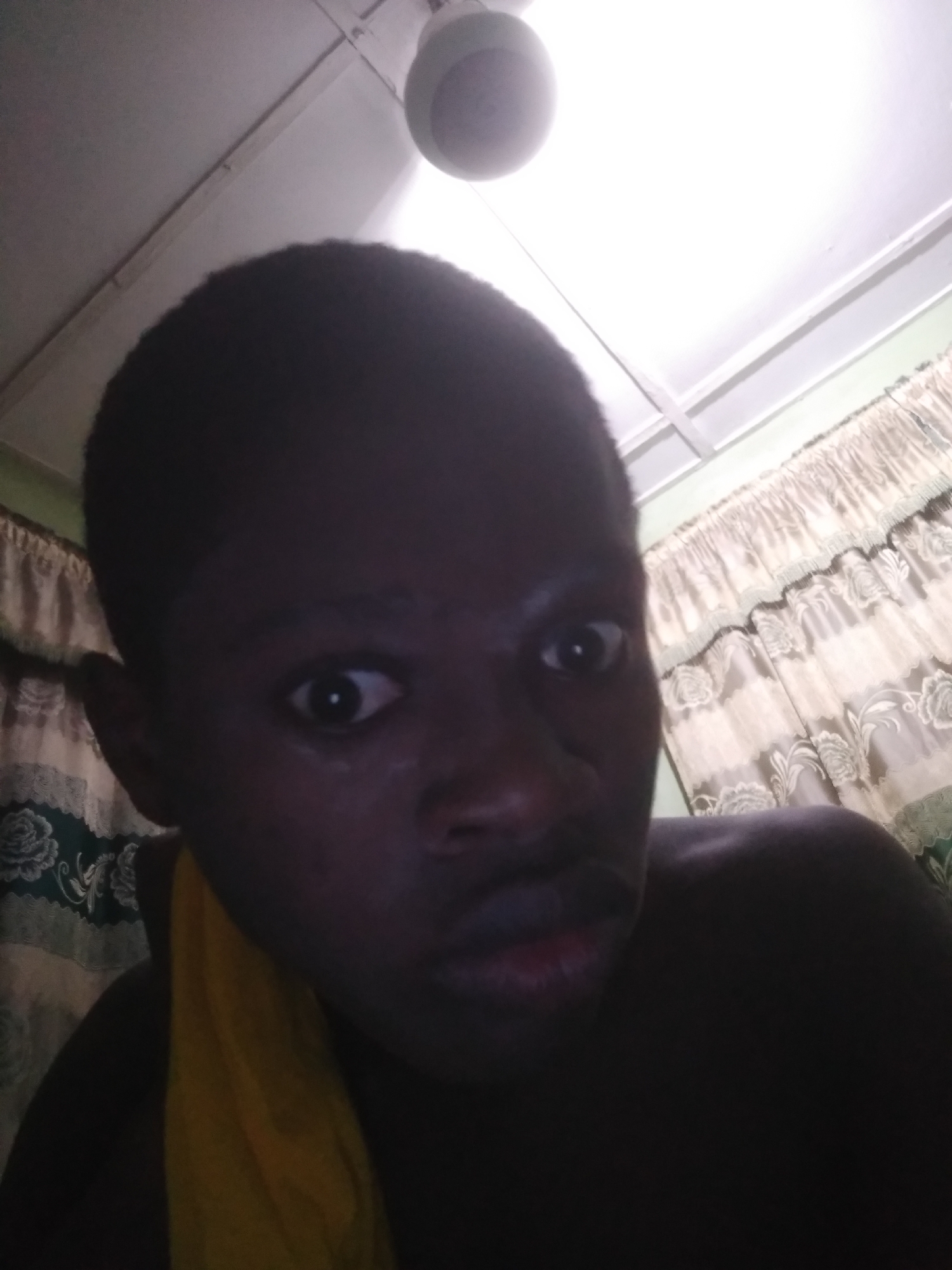 A person with dark skin is depicted in a close-up, low-angle shot. The person has short, dark hair, wide eyes, and is partially obscured by shadow. A yellow cloth or garment is draped over their shoulder. The background features a white ceiling with a light fixture and patterned curtains. The location is indoors, possibly a bedroom, in an unknown city.