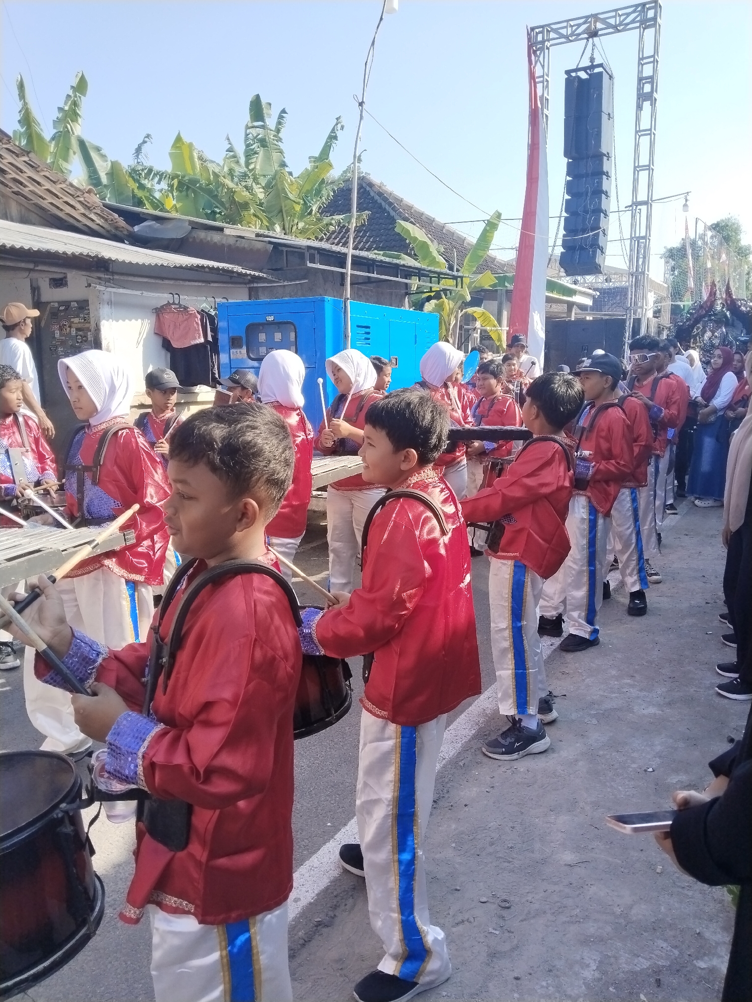 The image captures a street scene in what appears to be Gondang, Indonesia, likely during a parade or performance, given the presence of a marching band. A group of children, dressed in red jackets with gold trim and white pants with blue stripes, are lined up. Many of them are holding drumsticks, and some are positioned near drums or glockenspiels, suggesting they are part of a musical ensemble. The children are diverse in age, with some wearing white hijabs.
In the background, there's a large blue generator unit, a metal truss structure supporting a large speaker system, and a tall flagpole with Indonesian flags. Buildings with tiled roofs and vegetation, including banana trees, are visible behind the structures. A few adults are also present, some observing the children and others appearing to be participants or organizers. The ground is a paved, dusty surface. The sky is clear and blue, indicating a sunny day. The overall atmosphere suggests a community event or celebration.