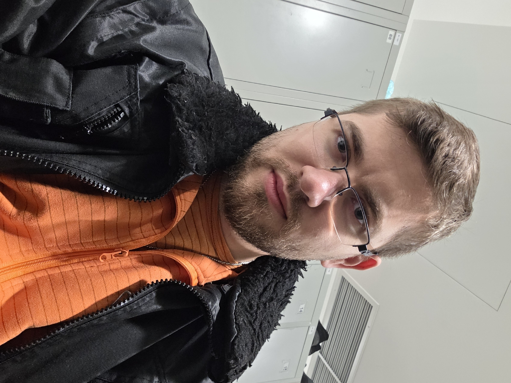 A selfie of a person with fair skin, brown hair, and a beard, wearing glasses, an orange striped shirt, and a black jacket with a furry collar. The person is indoors, with lockers visible in the background, potentially in a workplace or school setting. The location cannot be determined as the city of Plovdiv, Bulgaria, solely based on this image. The subject is facing the camera and is likely holding a phone or camera to take the selfie.