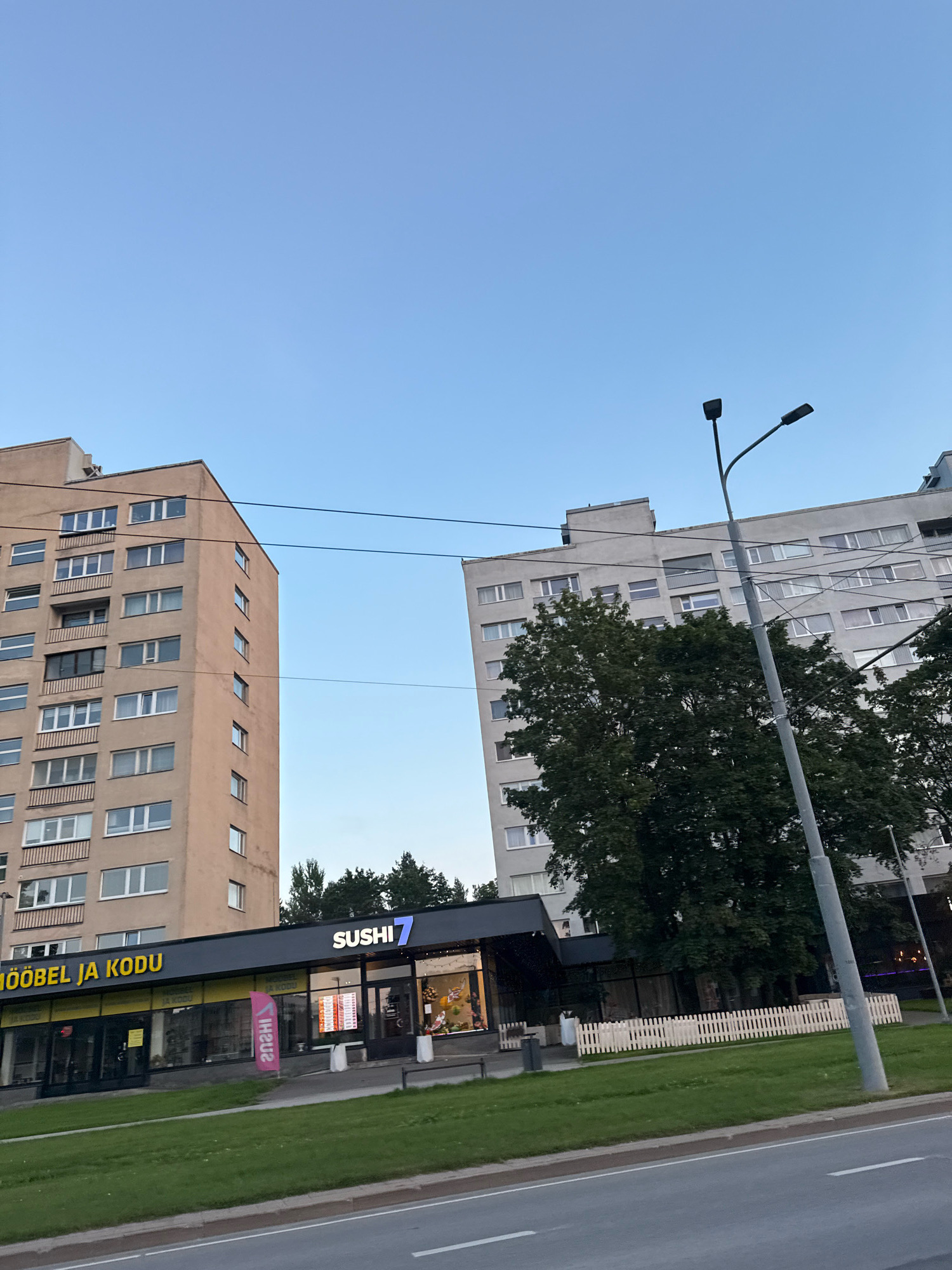 The image shows a street-level view of two apartment buildings in Tallinn, Estonia. The building on the left is a taller, beige-colored apartment block. To its right, a darker building with a similar facade is visible. In front of these buildings, at street level, is a commercial area. A sign with "MÖÖBEL JA KODU" is visible on the left side of the commercial area, indicating a furniture and home goods store. To the right of this is a "SUSHI 7" restaurant, recognizable by its illuminated sign. A white picket fence runs along the front of the commercial area. A light pole with two lamps stands prominently in the foreground, and power lines are visible crisscrossing the sky. Trees are situated between the buildings and the commercial area, and a grassy lawn is in front of the stores. The sky is a clear, pale blue, suggesting it is daytime.
