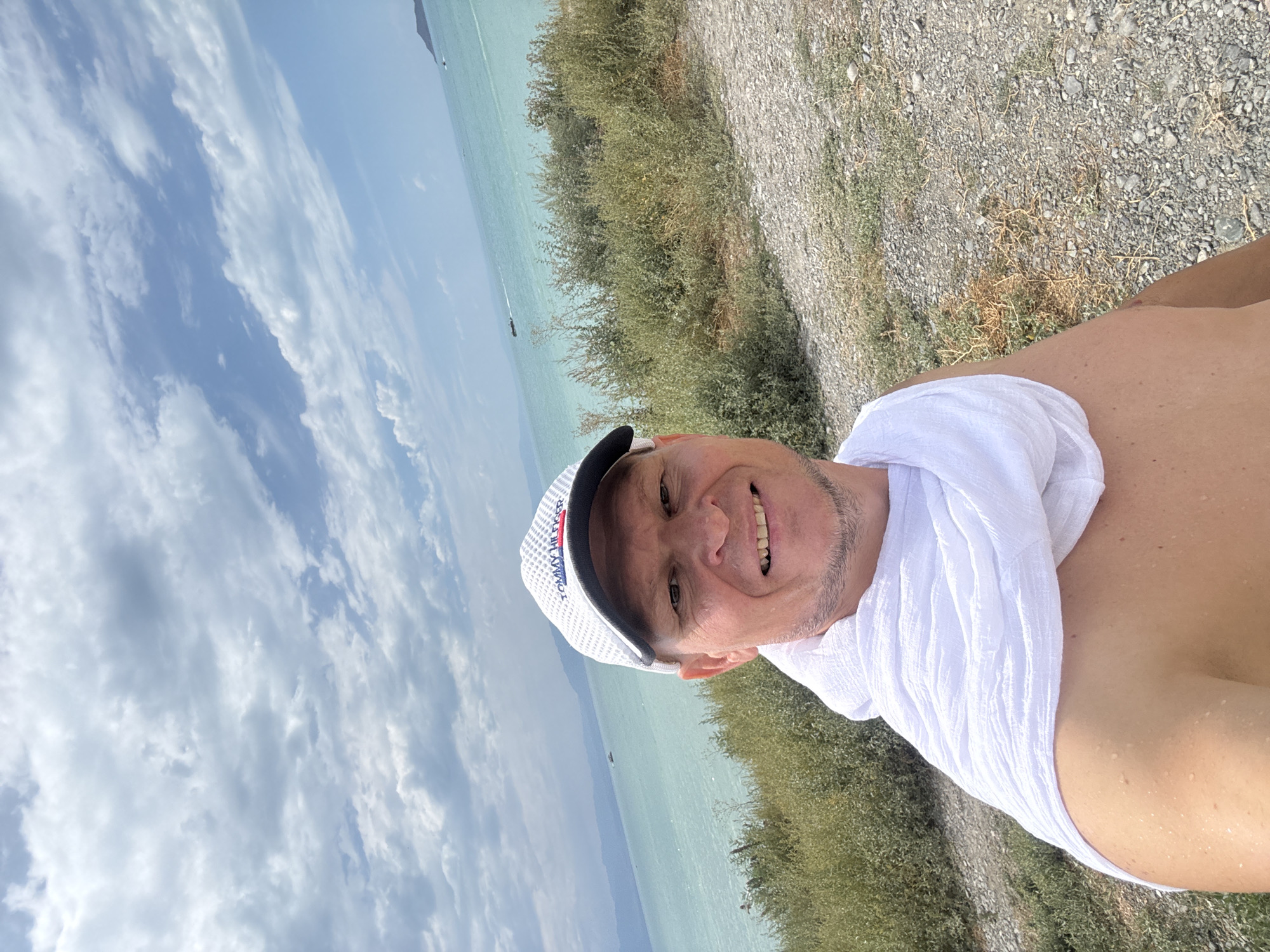 A man in a white baseball cap and a white scarf around his shoulders takes a selfie outdoors. He is smiling and his skin appears to be damp. Behind him, a vast body of turquoise water stretches to a hazy horizon. The water is dotted with a few boats. The landscape is covered with dry brush and gravel. The sky is filled with scattered white clouds. The man is standing on a slight incline, and the terrain slopes downwards towards the water. The location is likely near the city of Urdzhar, Kazakhstan, given the proximity to a large body of water.