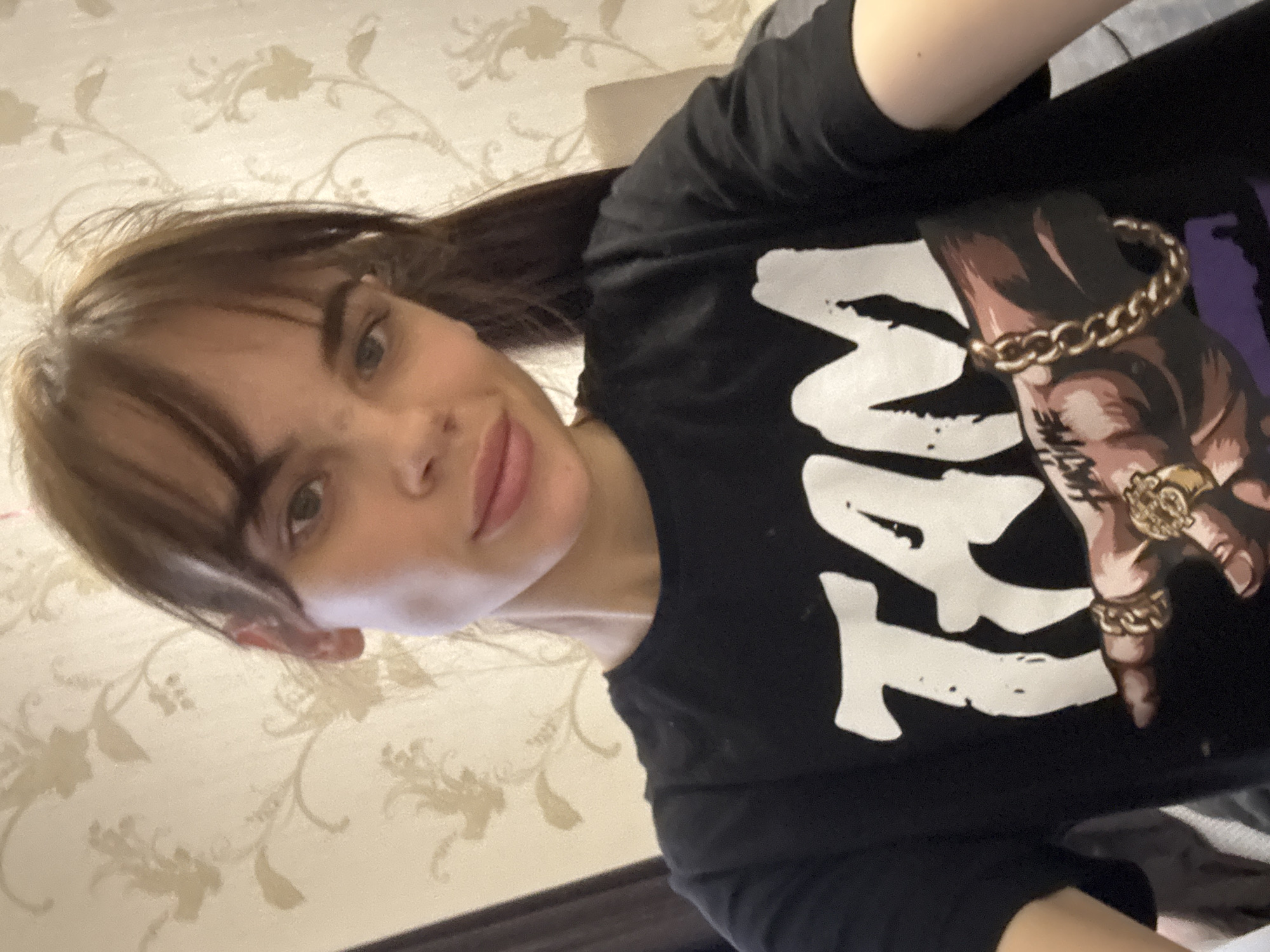 A young woman is posing for a selfie. She has brown hair with bangs and is wearing a black t-shirt with a graphic print of a hand wearing rings and a bracelet. The background is a wall with a floral pattern. The location is not discernible from the image.