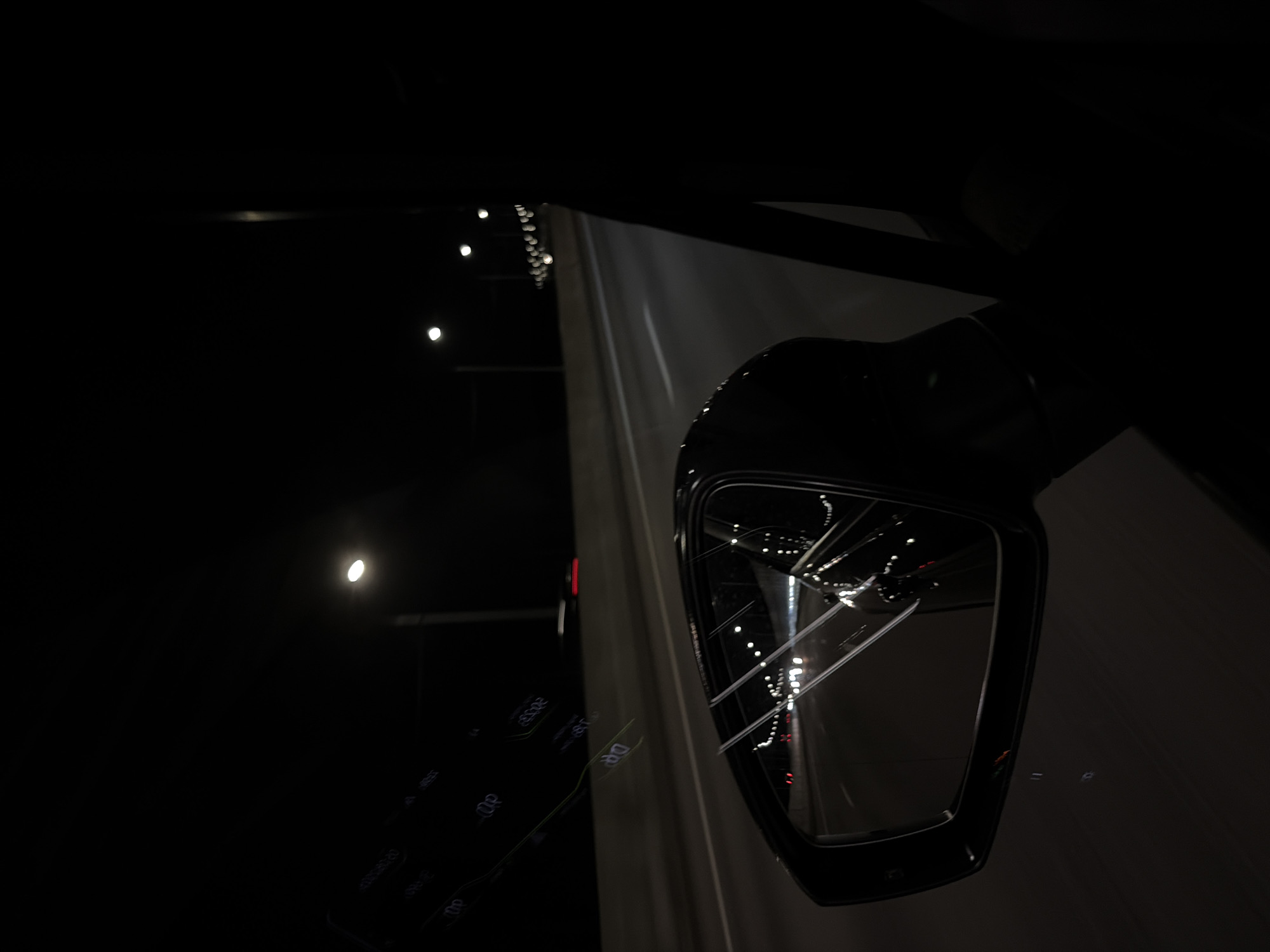 The image depicts a nighttime view from inside a moving vehicle, focusing on the side-view mirror. The mirror reflects the road ahead, which is illuminated by streetlights and the headlights of other vehicles. The road appears to be a multi-lane highway, with a concrete barrier separating the lanes. In the background, numerous streetlights are visible, stretching into the distance along the highway. The sky is dark, indicating it is nighttime. The interior of the car, including the door frame and window, are visible in silhouette. There are some faint reflections on the windshield that appear to be digital readouts. No people are directly visible, but the presence of a vehicle implies occupants. The location is specified as the city of Poltevo, Russia.