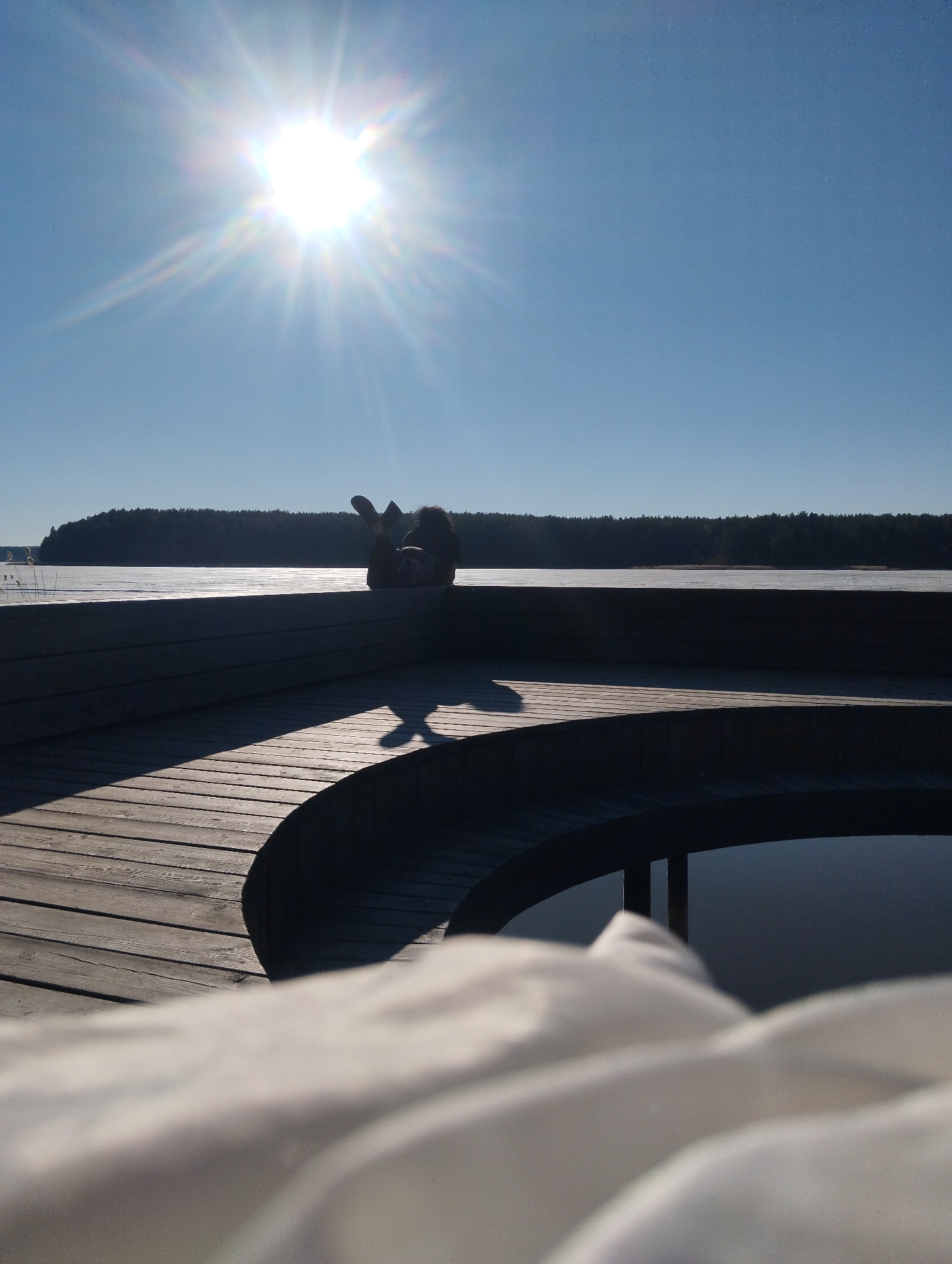 The photograph captures a scene on a wooden deck with a person lying down near the edge, overlooking a body of water that appears partially frozen. The setting is under a bright, clear sky with a strong sun providing intense light.
In the distance, a line of dark trees forms the horizon. The wooden deck is constructed with multiple levels, creating shadows across the boards. A curved section of the deck gives way to an opening, revealing the dark water below. A white, soft object, possibly bedding or clothing, is in the foreground.
The person lying down is silhouetted against the bright backdrop of the partially frozen water, making it difficult to discern specific details about them.