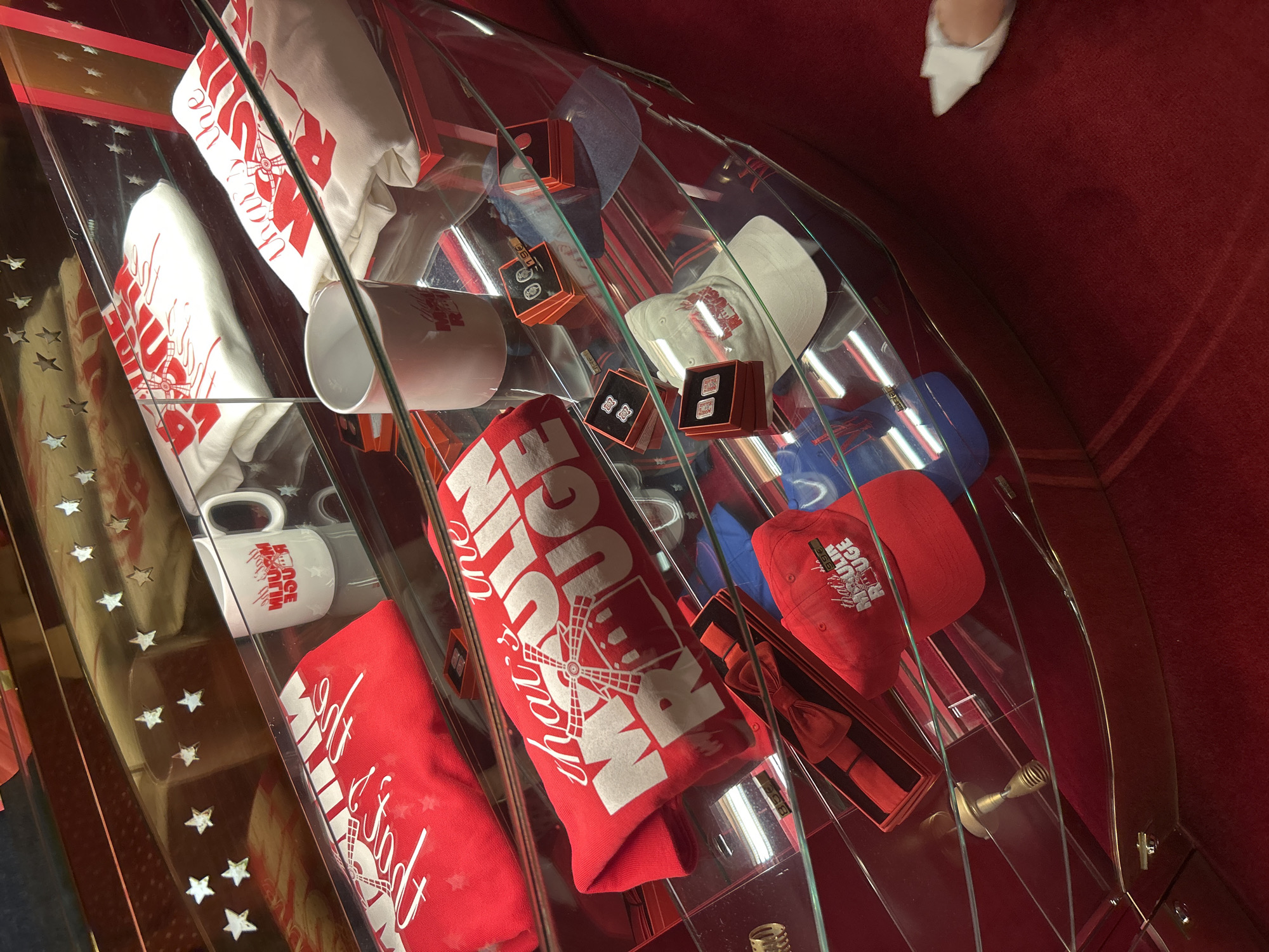 The image shows a display case containing merchandise themed around "Moulin Rouge." The display is illuminated and features multiple levels showcasing various items. Prominently displayed are red t-shirts and hats with white text and graphics, including a windmill, that read "that's the Moulin Rouge." There are also white mugs with similar red text. Smaller items like cufflinks or tie clips are presented in red boxes. A white baseball cap is visible, and a red bow tie is neatly presented in its packaging. The background of the display case is decorated with small gold stars on a red surface. The entire display rests on a plush red carpet. There is no indication of the city of Paris, France, within the image itself.