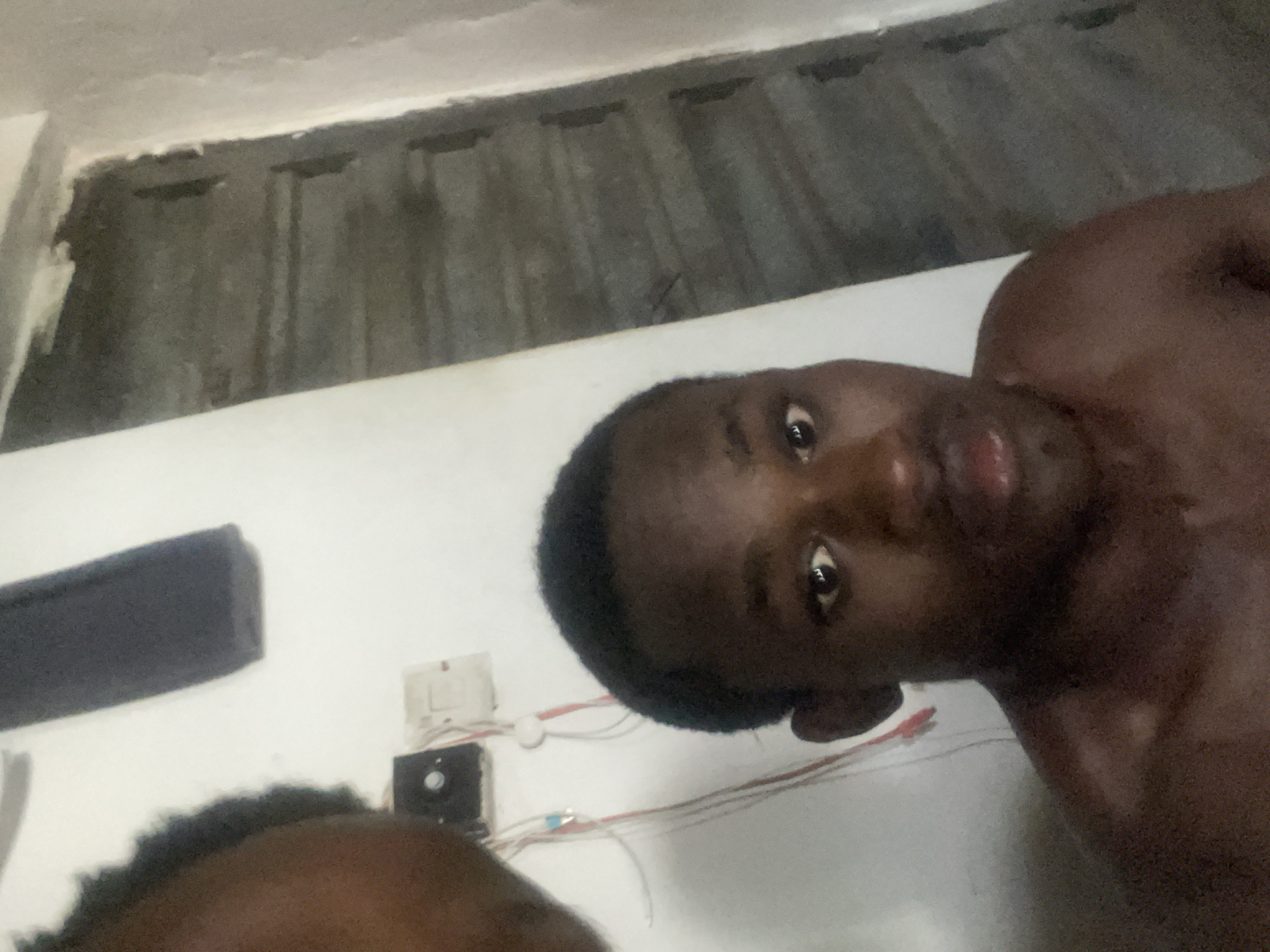 A medium shot selfie shows a Black male, shirtless, looking directly at the camera. He has short, dark hair and his face is partially visible on the right side of the frame. The background consists of a white wall with some electrical outlets and wires, and a darker, textured surface on the right, possibly a door or wall. The lighting is dim, creating shadows on the subject's face and body.