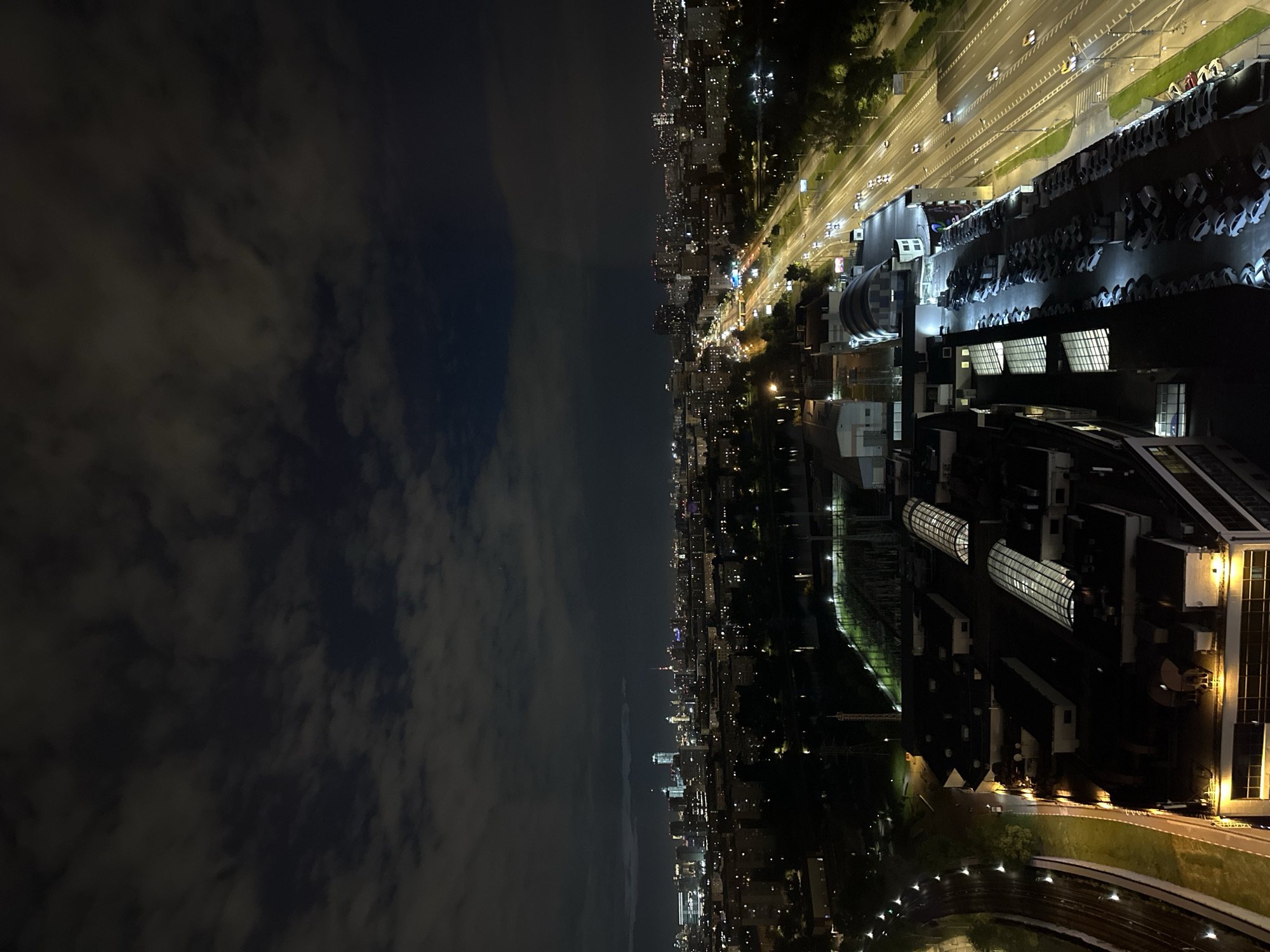 Captured from an elevated perspective, this nighttime photograph presents a cityscape. The scene is sharply bisected, with the left side dominated by a dark, cloudy sky and the right side depicting an illuminated urban environment. The cityscape extends into the distance, marked by the lights of buildings and streets.
A prominent feature is a large building complex with modern architectural elements, positioned to the right in the foreground. Adjacent to this building is a substantial parking area densely populated with vehicles. A highway runs parallel, with the streaks of headlights and taillights indicating active traffic. In the distance, the skyline of Moscow is visible, with the subtle glow of city lights blending into the night sky. The overall environment is quiet and still, illuminated by artificial light sources.