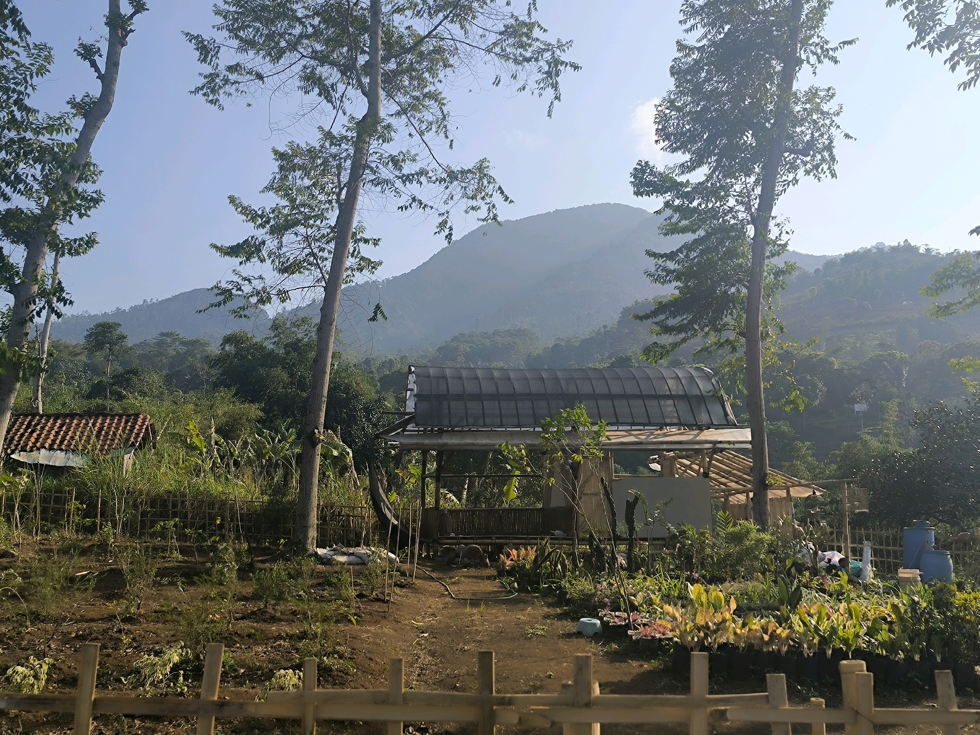 The image shows a rural mountainous landscape near Bandung City, Indonesia. In the foreground, a wooden fence borders a patch of dry earth with sparse vegetation and small saplings. A path leads to a simple, open-air structure with a dark, translucent roof and bamboo walls, possibly a pavilion or covered seating area. To the left, a small building with a terracotta tiled roof is visible, partially obscured by greenery. The midground is densely populated with trees and lush vegetation. In the background, rolling hills covered in trees rise to meet a hazy, light blue sky. The overall scene suggests a tranquil, natural environment, likely a farm or a natural reserve. There are no visible people in the image.