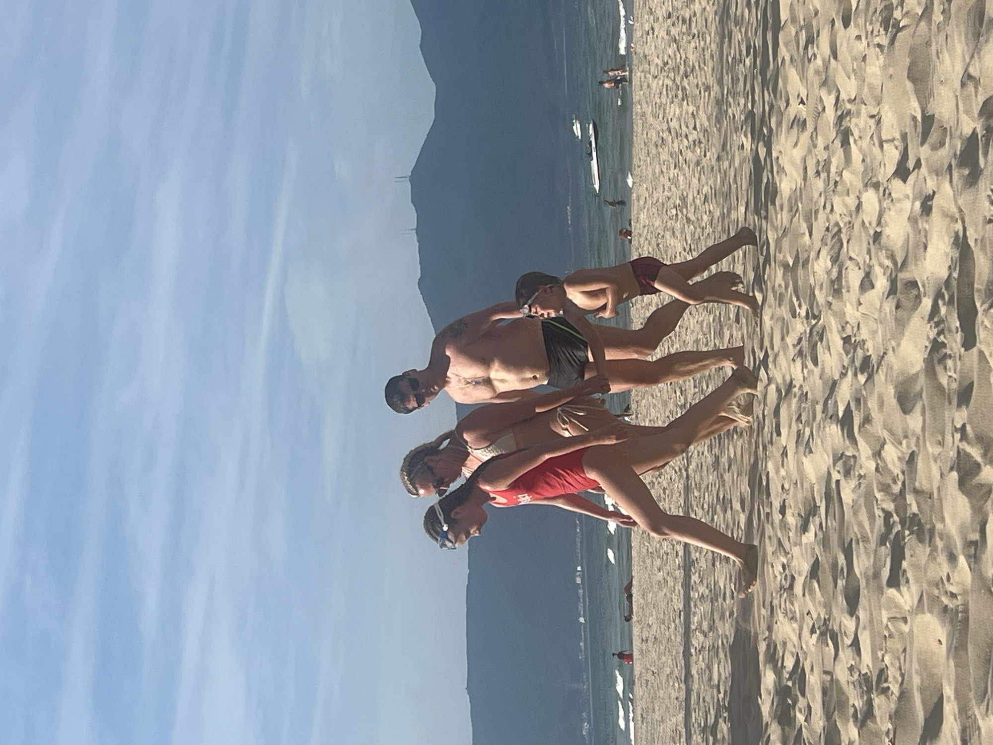 A family of four, two adults and two children, are walking on a sandy beach in Da Nang, Vietnam. The father, wearing black swim trunks and sunglasses, walks to the right of the frame. Next to him, a young boy in maroon swim trunks runs ahead, looking excited. To the father's left, a woman in a white bikini walks, with a young girl in a red swimsuit and goggles to her left, who is looking down. In the background, the ocean stretches out with gentle waves and a few boats. The sky is a clear blue with some wispy clouds, and a range of dark mountains forms the horizon. The beach itself is composed of light brown sand with ripples and shadows.