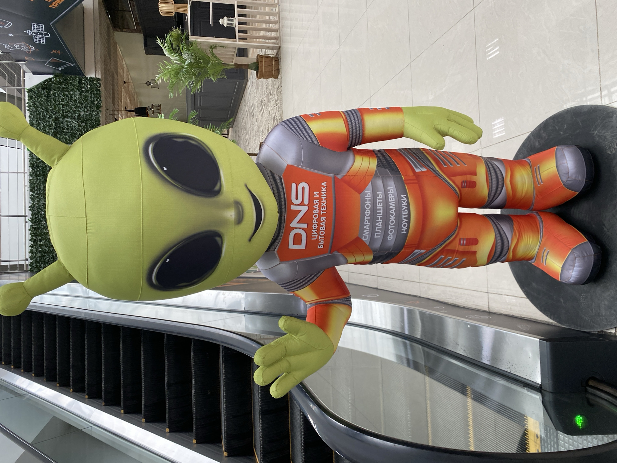An inflatable alien mascot stands next to an escalator. The alien is predominantly green with large black eyes and two antennae. It is wearing a silver and orange suit with the logo "DNS" and text in Russian indicating "digital and home appliances." The alien is waving with its left hand. The escalator is black with silver railings and has a green light illuminated at its base. In the background, there is a wall covered in greenery, a counter, some potted plants, and lighting fixtures. The floor is tiled. The location is identified as the city of Oral, Kazakhstan, likely within a shopping center or commercial establishment.