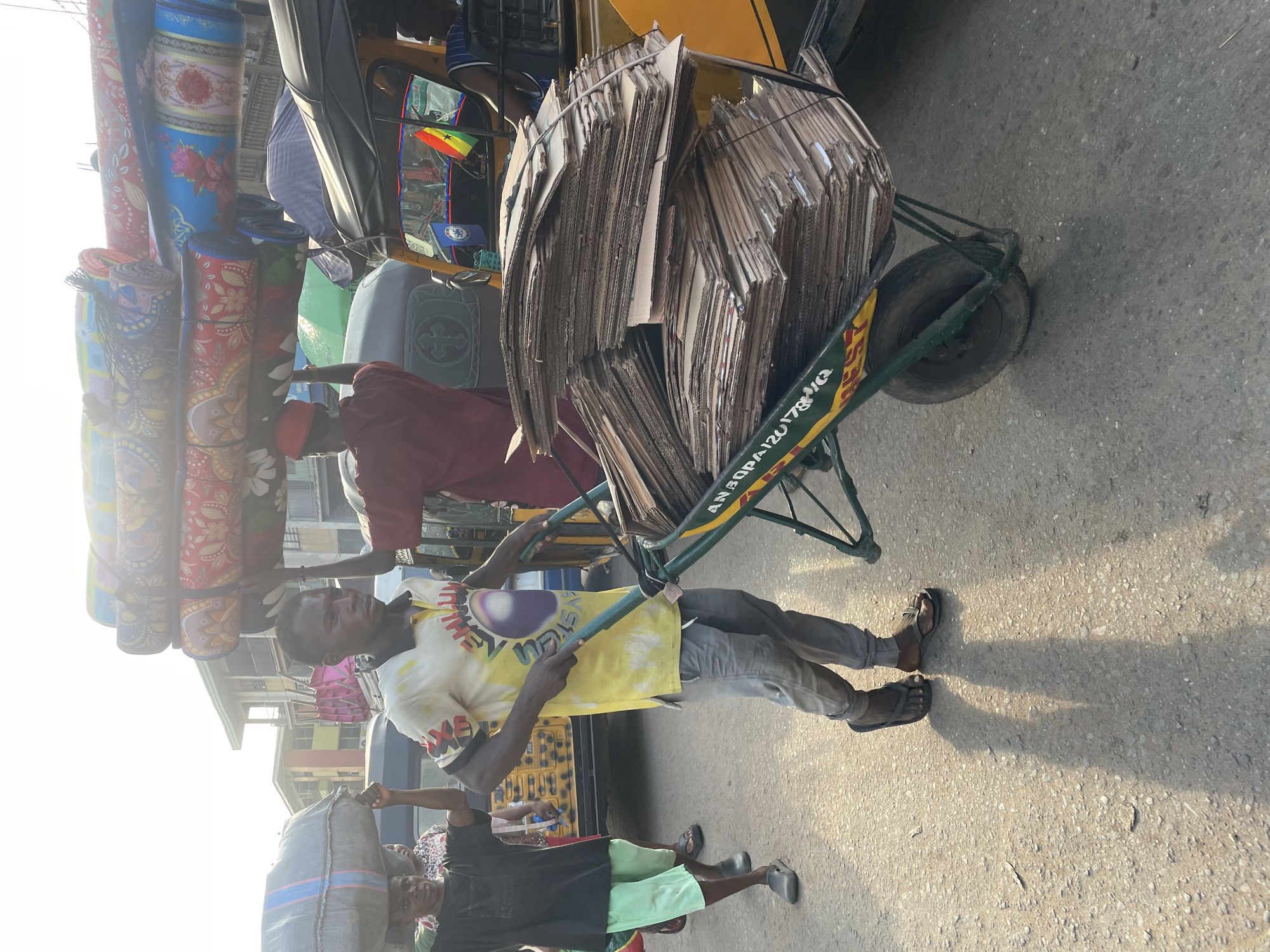 In Nkpor, Nigeria, a man pushes a wheelbarrow loaded with a large stack of flattened cardboard boxes down a sunlit street. The man wears a yellow t-shirt with colorful graphics and gray pants, and appears to be walking alongside a yellow motorized rickshaw. In the background, other individuals are seen carrying bundles of fabric rolls on their heads, and a child is visible near the front. The environment suggests a bustling market or roadside commerce area with buildings and vehicles present.