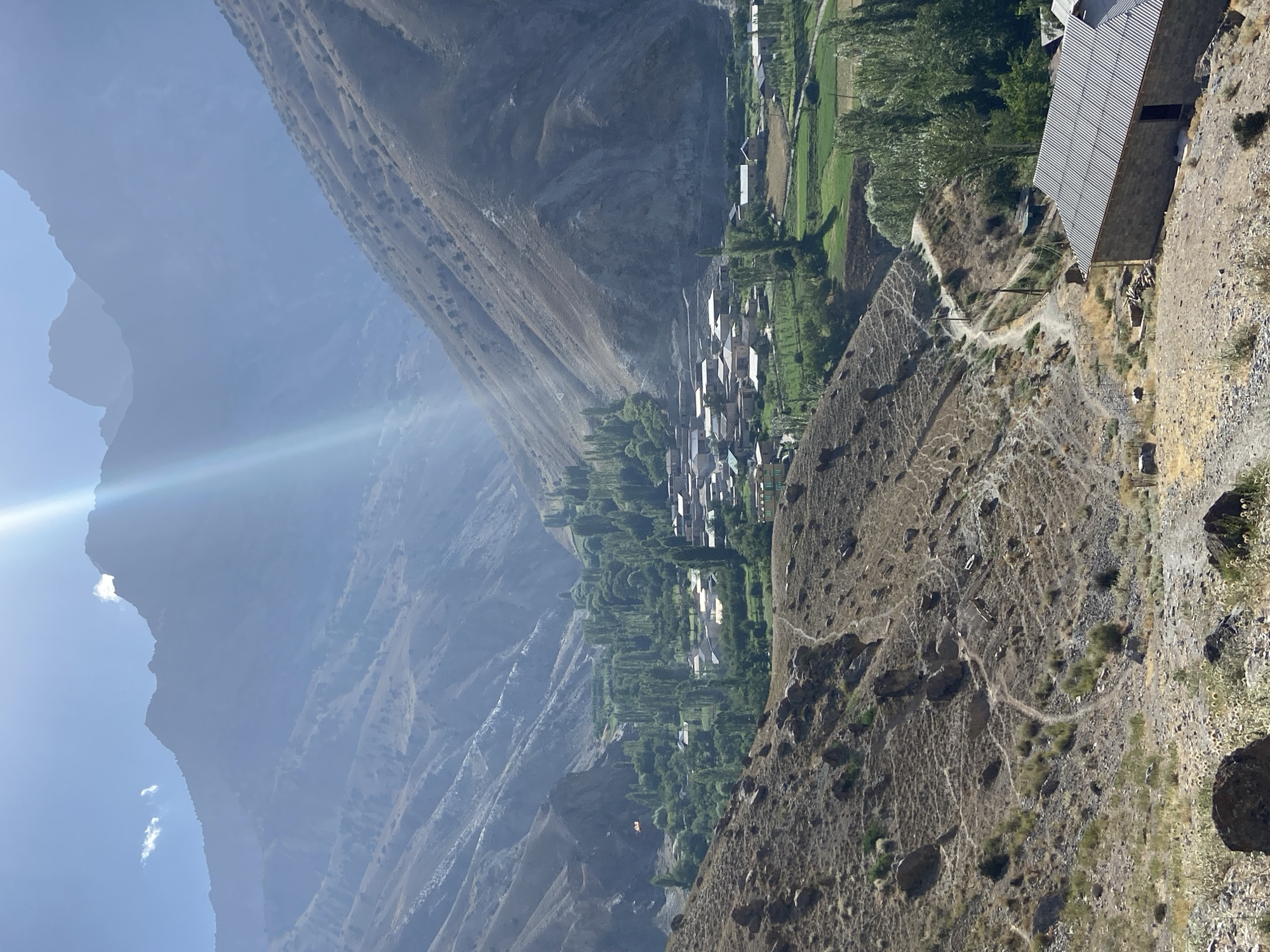 The image shows a rural village nestled within a valley in the Pamir Mountains, likely in the vicinity of Ghazza, Tajikistan. In the foreground, a barren, rocky hillside descends into the valley, with a dirt path winding down. A stone building with a corrugated metal roof is visible in the lower right corner. The valley floor is covered with lush green fields and a cluster of buildings, indicative of a village. Tall, slender trees, characteristic of poplar varieties, are scattered throughout the village and line the edges of the fields. The surrounding mountains are steep and rugged, with slopes showing signs of erosion and sparse vegetation. The sky is bright blue, with a strong sun casting a lens flare across the upper portion of the image, highlighting the haze and atmospheric conditions typical of high-altitude regions. The scene conveys a sense of remote, natural beauty and traditional rural life.