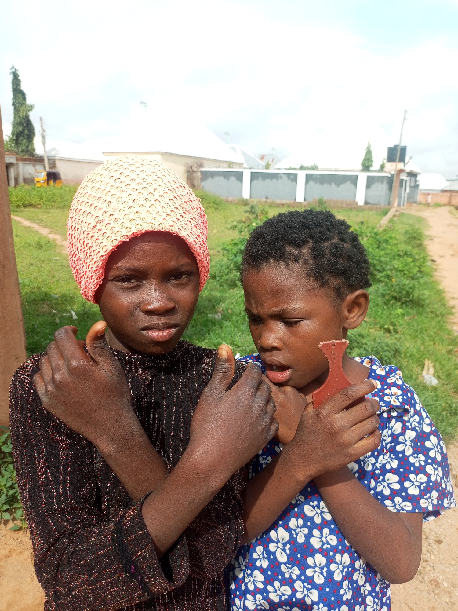 Two young girls are standing outdoors in what appears to be a residential area of Tempure, Nigeria. The girl on the left wears a textured, peach-colored head covering and a black shirt with metallic stripes. Her arms are crossed in front of her chest. The girl on the right has dark, short, curly hair and wears a blue shirt with a white floral pattern. Her mouth is slightly open, and she is holding a reddish-brown object, possibly a toy or tool, which is partially obscured by her hands and the other girl's arms. The background shows green vegetation, a dirt path, some buildings with white roofs and grey walls, a tall tree, and a utility pole. The sky is cloudy and bright.