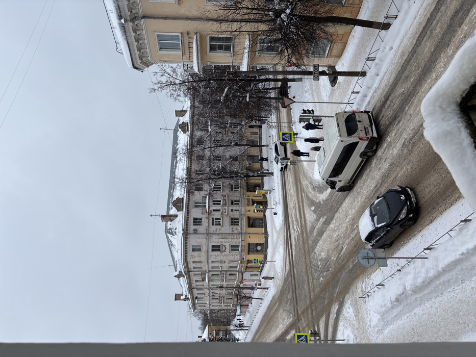 The scene depicts a snow-covered urban intersection in Velikiye Luki, Russia, specifically at the corner of Komsomolskaya Ulitsa and Ulitsa Karla Libknekhta, viewed from an elevated position, likely a building balcony. The sky is overcast and grey.
The ground is blanketed in snow. Roadways are partially cleared, revealing wet, dark asphalt and slush, with visible tire tracks. Sidewalks and unplowed areas are covered in a deeper layer of white snow. Bare deciduous trees with snow clinging to their branches are interspersed throughout the environment.
In the midground-left, a multi-story (at least four floors) light-colored building, primarily off-white or pale beige, with yellow-painted commercial facades on its ground floor, forms the prominent corner of the intersection, identified as Komsomolskaya Ulitsa 29 / Ulitsa Karla Libknekhta 12. Its roof and ledges are covered in snow, and it features classical architectural elements including cornices and balconies. On the right, a section of a similar light-yellow building, likely Komsomolskaya Ulitsa 31, is visible with snow and icicles on its roof and decorative moldings.
Vehicle activity includes a black Peugeot 308 sedan, with snow accumulated on its roof and hood, traversing the foreground from left to right. Its Russian license plate is partially visible. A white commercial van or minibus is positioned in the midground, also moving left to right, near a group of pedestrians. A white SUV is stationary at a crosswalk further up the street on the left.
Four pedestrians, dressed in winter clothing, are standing on the snow-covered sidewalk in the midground-right, adjacent to a traffic light and a pedestrian crossing, appearing to wait to cross the street. Additional individuals are visible walking on sidewalks in the background.
Visible road signs include multiple blue-and-white pedestrian crossing signs, a red-and-white "СТОП" (STOP) sign mounted on a pole, and a "Give Way" sign. Operational traffic lights are also present. The immediate foreground shows a snow-covered balcony railing, indicating the elevated viewpoint. Overhead power lines span the sky.