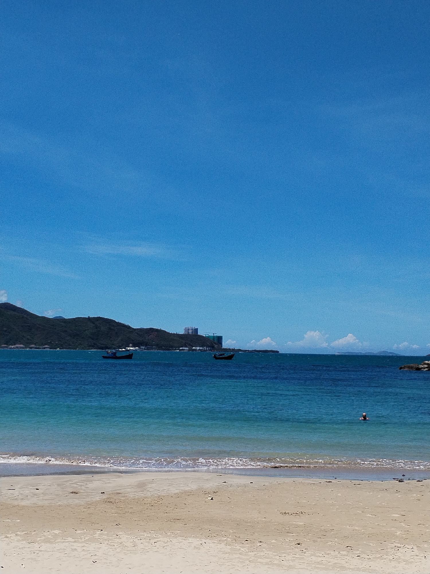The image shows a coastal scene in Bac Nha Trang Ward, Vietnam. A sandy beach occupies the foreground. The water transitions from shallow turquoise to a deeper blue, with small waves near the shore. In the distance, there's a hilly landscape. Three boats are visible on the water, and a person swims in the water closer to the beach. The sky is clear blue with some light clouds.