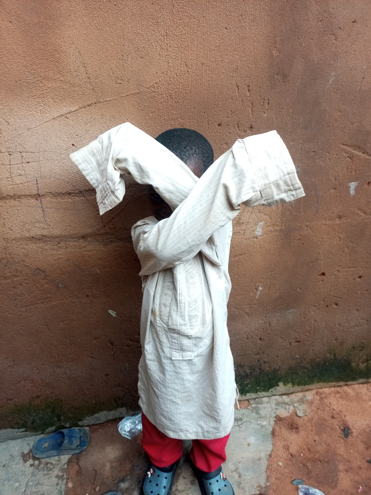 A child stands in front of a textured brown wall, with their arms crossed over their face, obscuring their features. They are wearing a light-colored, striped tunic over red pants and dark blue clogs with a floral detail. To the left of the child, a blue sandal lies on the ground. The ground is a mixture of concrete and dirt. The wall behind the child has some faint markings and a patchy, uneven texture. Green moss or algae is visible along the base of the wall. The scene appears to be an outdoor setting, possibly a residential area in Tempure, Nigeria.