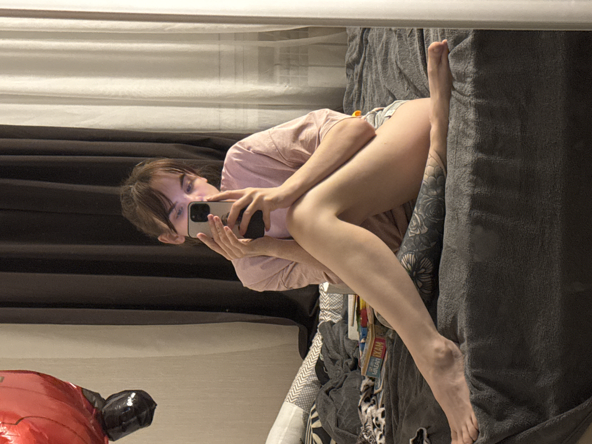 A woman is visible in a reflection, holding a mobile phone and facing it. She is wearing a pink shirt and shorts. Part of her leg and feet are visible next to a grey blanket. The background includes a curtain, a window blind, and what appears to be a red foil balloon. The location is unknown.