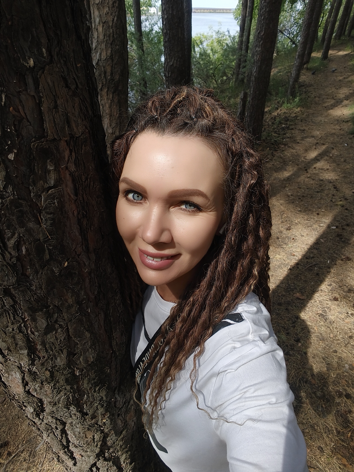 A medium shot captures a woman leaning against a tree. She is fair-skinned with blue eyes and her hair is styled in brown dreadlocks. She wears a white long-sleeved shirt and a black strap across her torso. The tree she leans against has rough, dark bark. The background shows other trees, a body of water, and a ground covered in dry grass and pine needles. The location appears to be a forest or park.