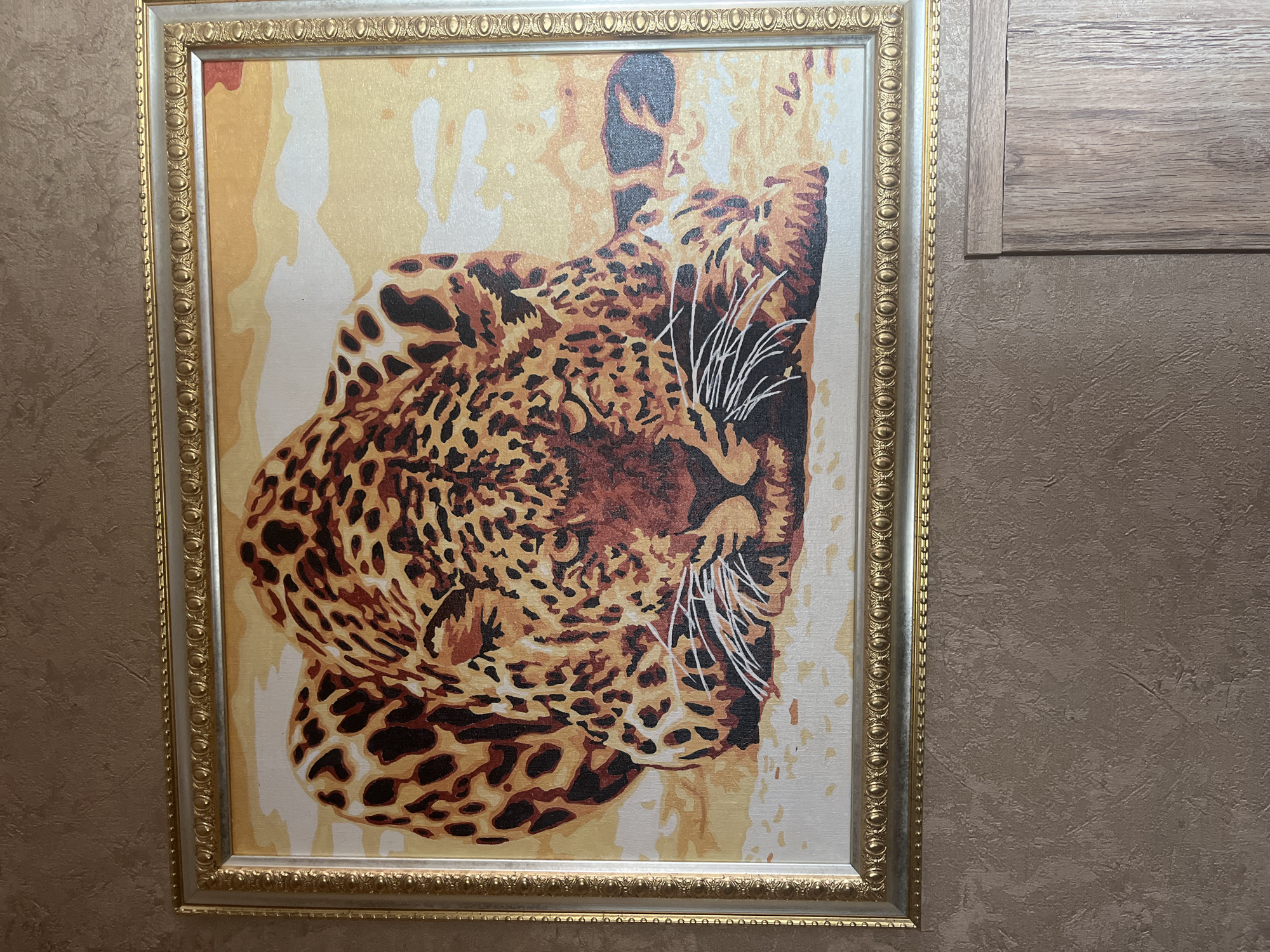 A framed painting of a leopard is displayed on a textured brown wall. The painting depicts a leopard lying down, with its head facing the viewer. The leopard has a distinctive spotted coat in shades of orange, brown, and black. Its eyes are alert, and its white whiskers are prominent. The background of the painting is a gradient of yellow and white, suggesting a bright, possibly sunlit, environment. The frame surrounding the painting is ornate, with a silver outer layer and a gold inner layer featuring a decorative pattern. The wall the painting is hung on has a rough, textured finish. To the right of the painting, a section of wooden paneling is visible. The scene appears to be an interior setting.