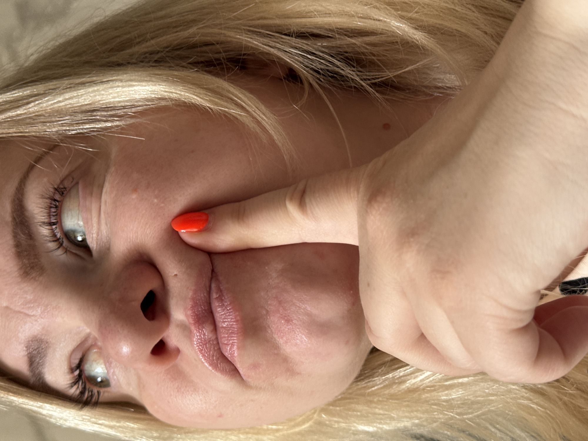 A close-up, rotated portrait captures a fair-skinned individual, likely female, with long blonde hair and light-colored eyes. The subject is positioned so their head is angled, possibly lying down. A hand with bright red nail polish on the index finger presses firmly into the person's left cheek, causing a noticeable distortion of the facial features, including the cheek, nose, and mouth. The lips are slightly parted due to the pressure. Both eyes are open, with prominent eyelashes, and are looking towards the upper left of the frame. The blonde hair cascades around the face, with individual strands visible. The skin exhibits natural texture, and a small, reddish mole or blemish is visible near the hairline on the temple. The background is out of focus, appearing to be composed of the subject's hair and possibly a soft surface. The lighting is soft and even, suggesting an indoor setting. The image depicts a moment of playful or experimental facial manipulation. No text is visible in the image. The location context for this media file is Chaikovsky, Russia.