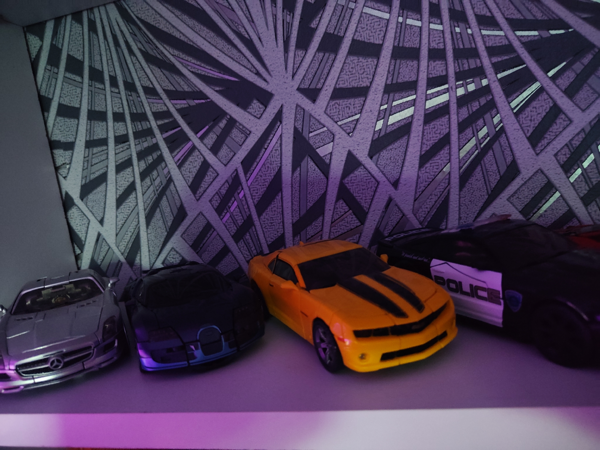 The image displays a collection of toy cars lined up on a white shelf against a backdrop with a geometric, angular pattern illuminated with purple and blue light. From left to right, the cars are: a silver Mercedes-Benz toy car, a dark blue toy car with glowing blue headlights, a bright yellow toy car with black racing stripes, and a black toy police car with "POLICE" written on its side. The lighting casts a purple hue on the shelf and the lower portions of the cars. The scene is indoors, and there is no information available about the city of Borova, Ukraine.