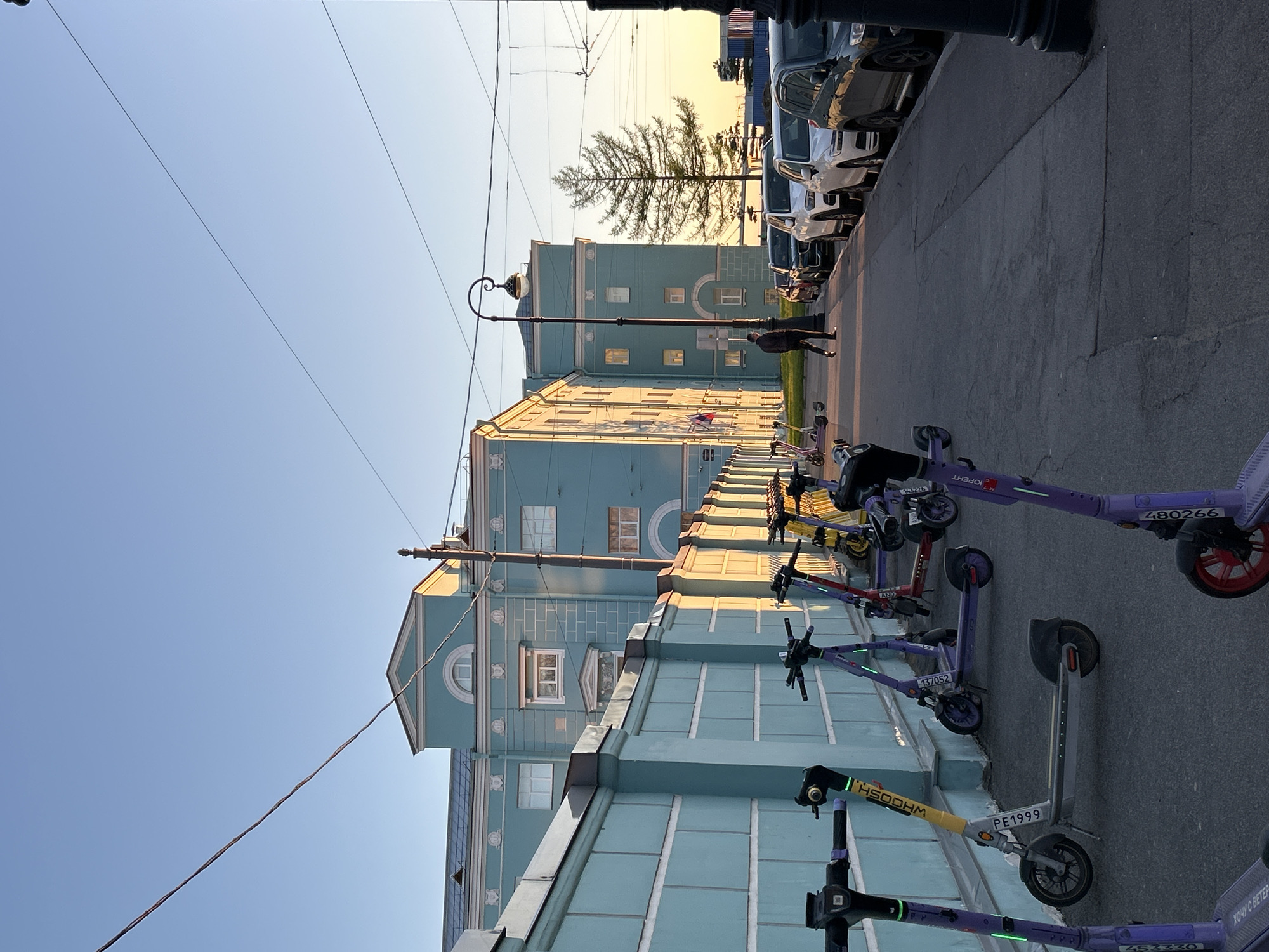 The image depicts a street scene in Saint Petersburg, Russia. The street runs along a light blue building and is lined with parked cars on the right side. A row of electric scooters is parked on the pavement next to the building. A lamppost stands in front of the building. Overhead electrical wires are visible against a blue sky with a bright yellow sunset on the horizon. A person is walking on the street.