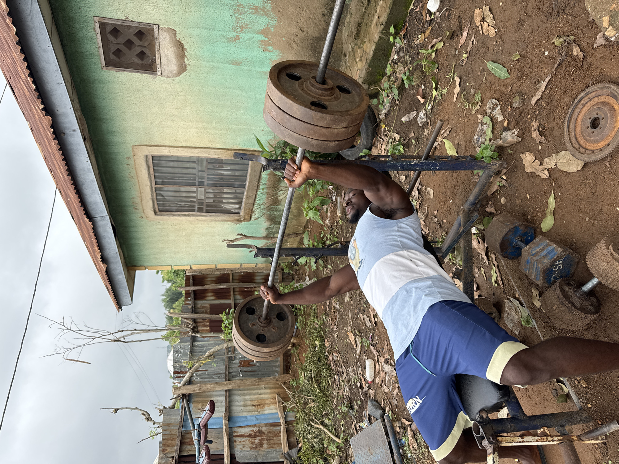 The image depicts a man lifting a barbell with weights outdoors in Akwanga, Nigeria. He is lying on a weight bench with a barbell in his hands, lifting weights. The surroundings include a house with a green wall and a rusty roof, a corrugated metal fence, dumbbells, a rusty metal disc, and foliage growing in the dirt. The sky is overcast.