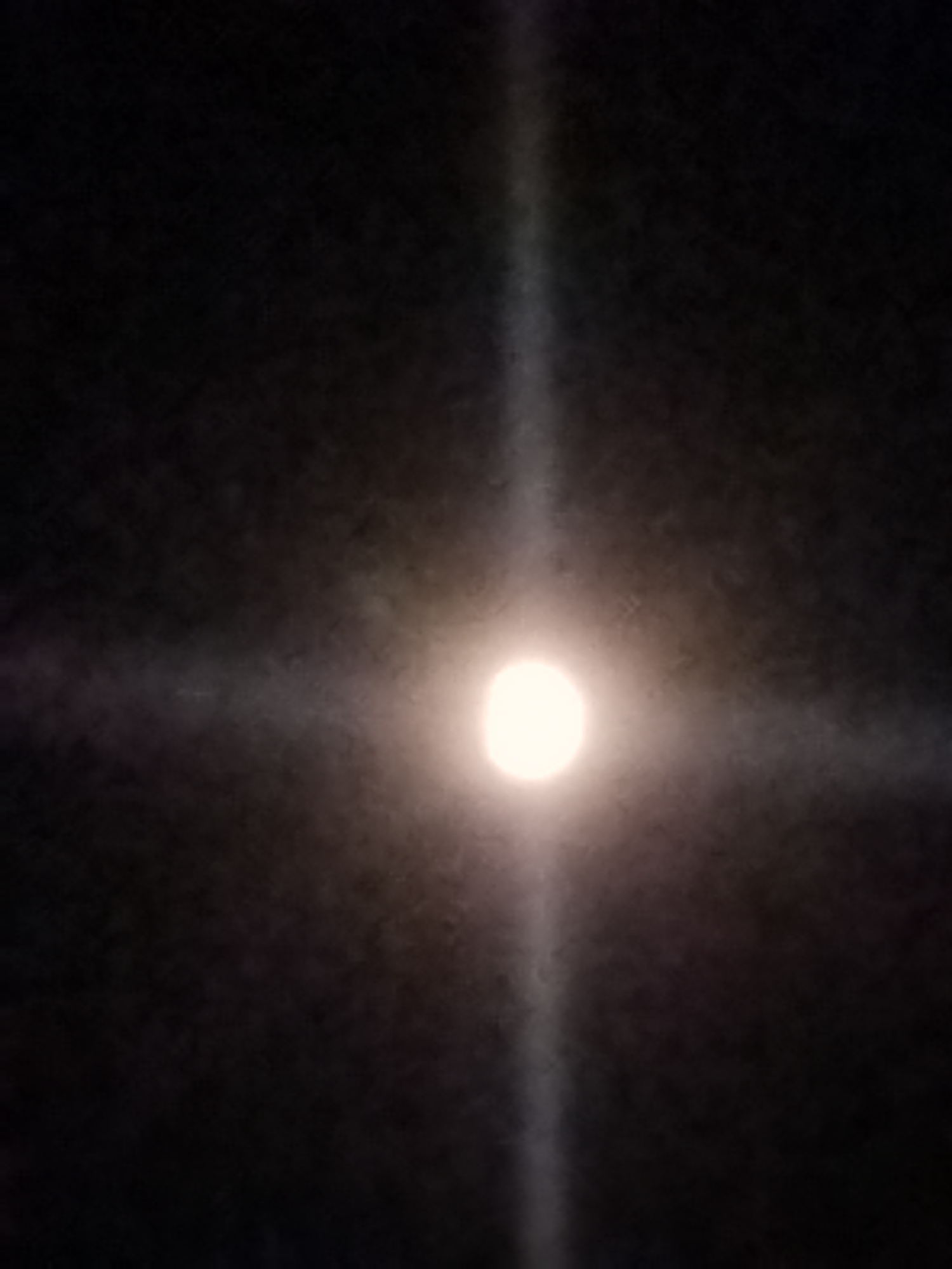 The image shows a bright, luminous circle in the center of a dark sky, resembling the moon or a distant light source. A distinct cross-shaped illumination emanates from this central point, extending outwards into the darkness. The light is diffused, suggesting it might be passing through atmospheric conditions like fog or dust. The overall impression is of a celestial event or a powerful light piercing the night. There are no discernible people, objects, or specific location cues within the frame that would indicate a particular setting beyond the dark sky. The time of day is clearly night due to the darkness. The weather is not definitively discernible, though the diffusion of light could suggest overcast conditions or the presence of moisture. No text is visible in the image.
